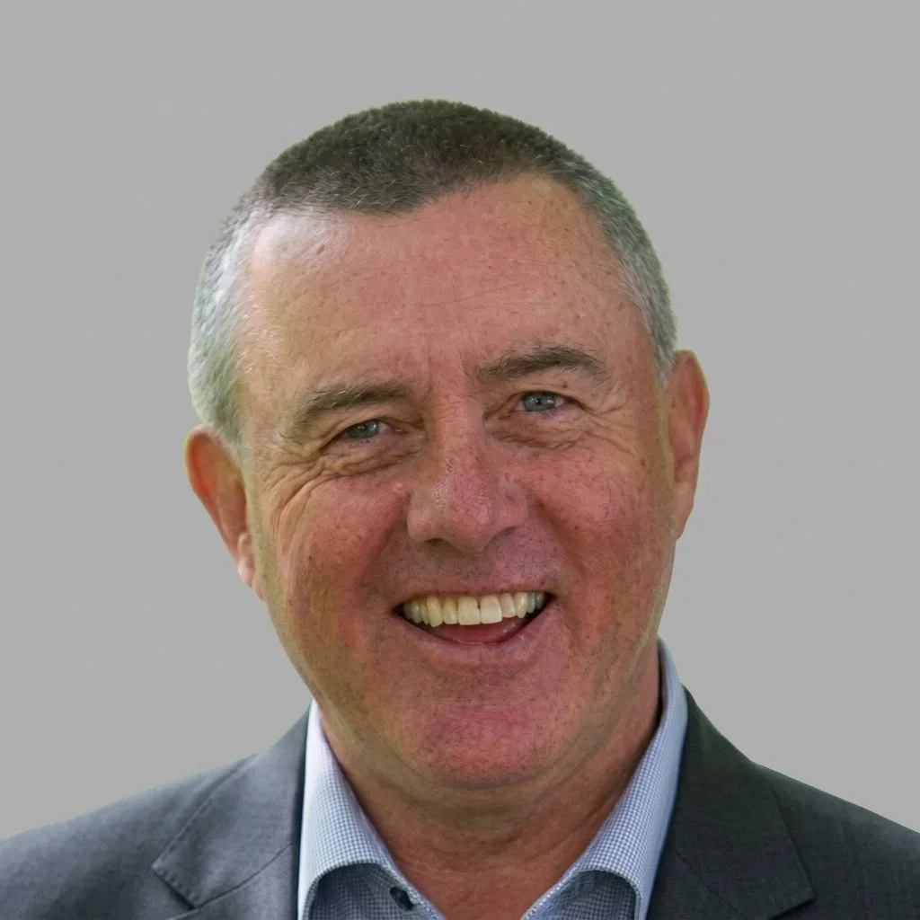 Smiling man with short hair outdoors, green trees in the background.