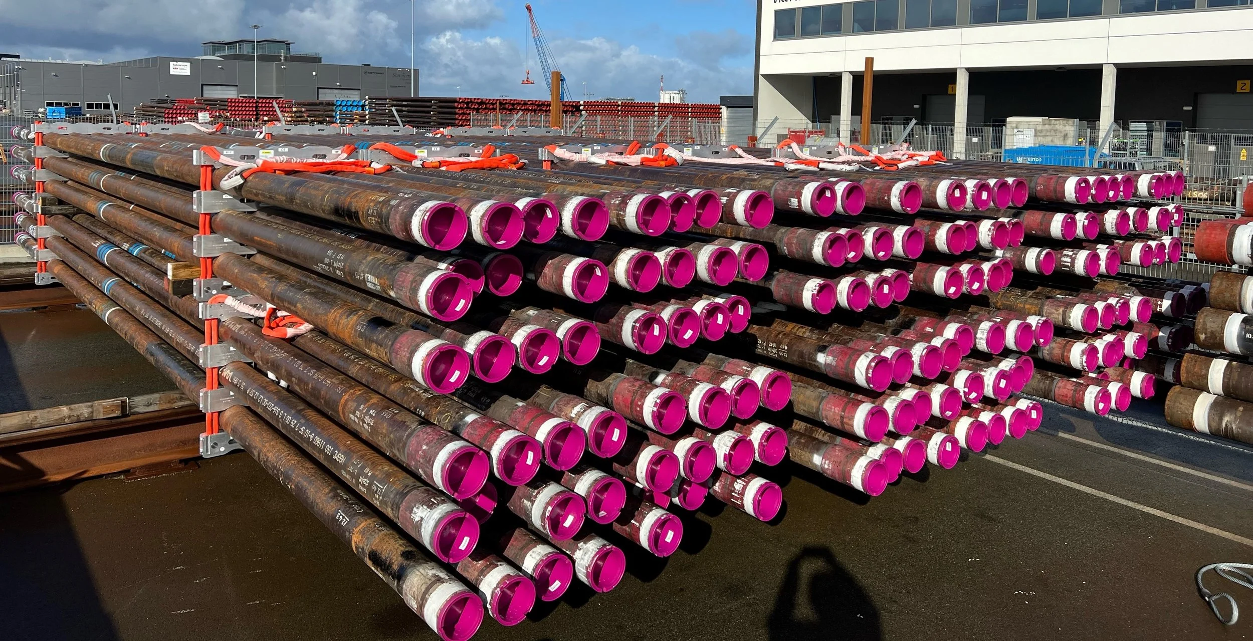 Stacked steel pipes with pink caps stored on a construction site outside with a building and crane in the background.