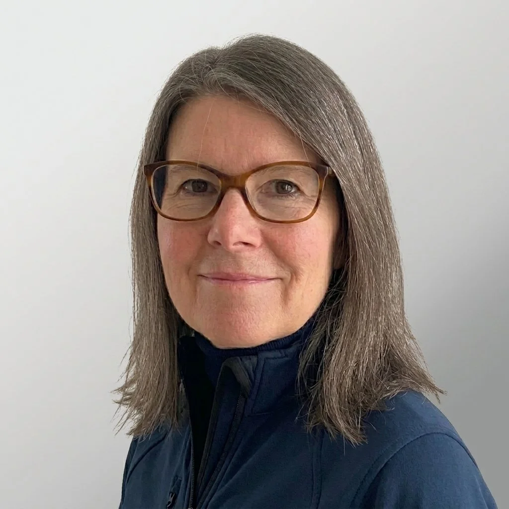 A woman with shoulder-length gray hair and glasses, wearing a black turtleneck and a dark blue jacket, posing against a plain light-colored wall.