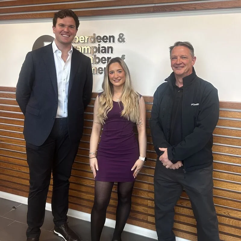 Three people standing indoors in front of a wooden wall with a sign, smiling at the camera. The person on the left is a tall man in a dark suit and white shirt. The person in the middle is a woman with long blonde hair in a purple dress and black tights. The person on the right is an older man in a black jacket and pants.