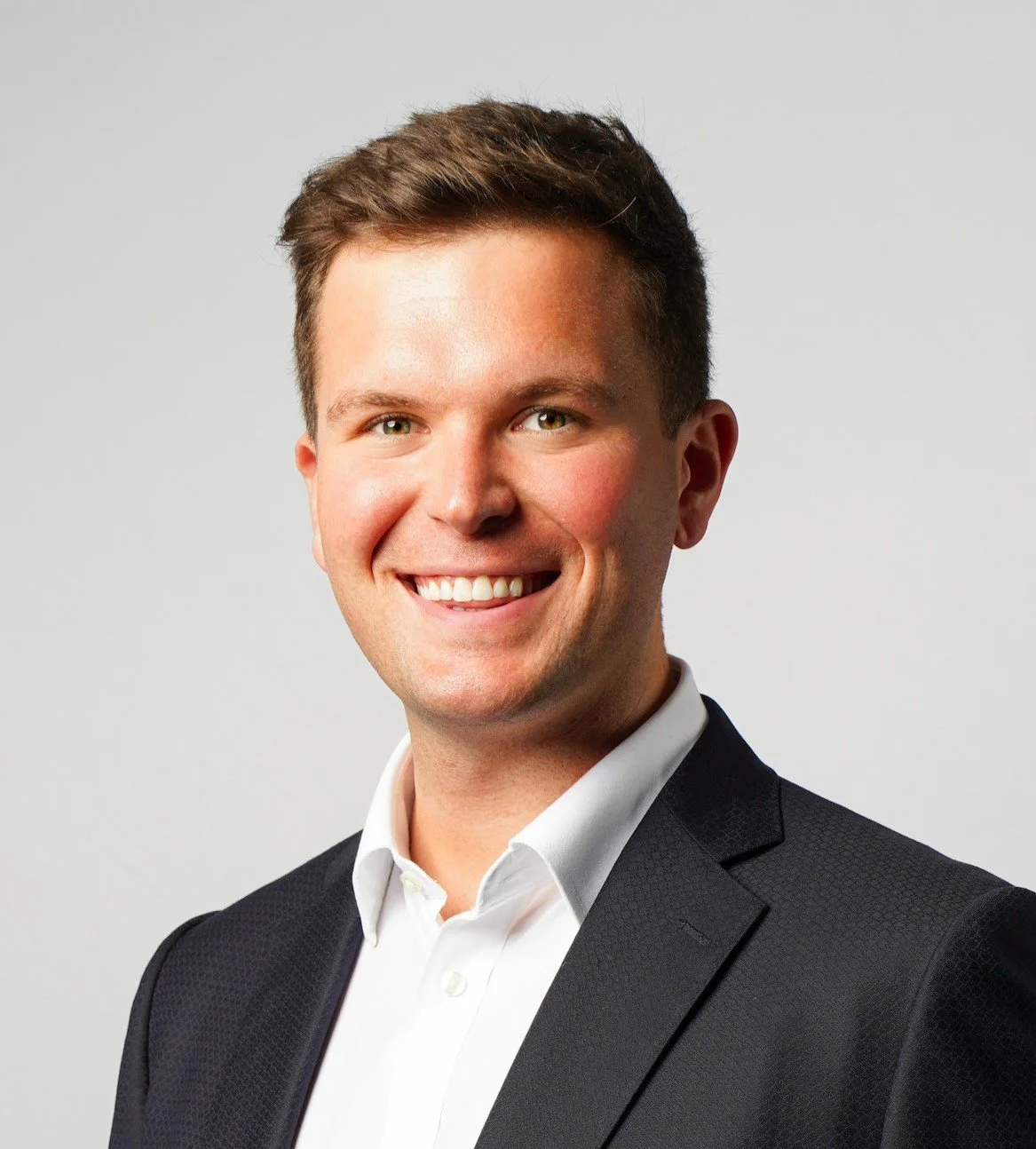 Professional headshot of a young man with short brown hair, smiling, wearing a white shirt and a black blazer, against a plain light gray background.