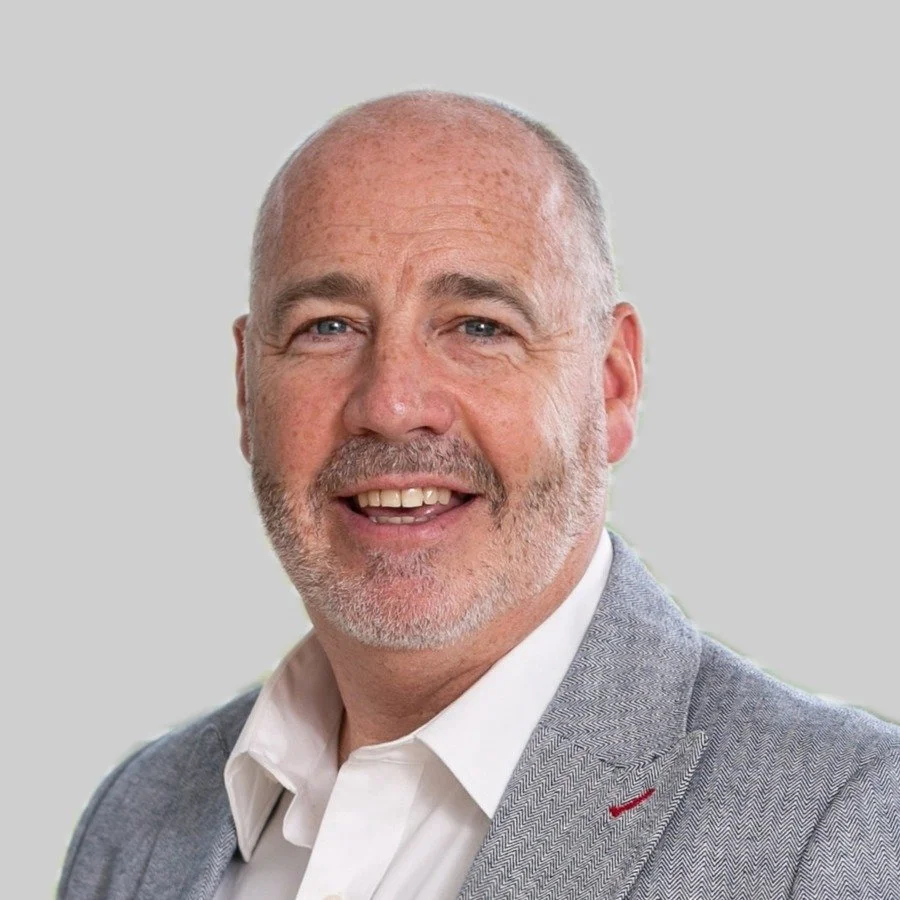 A middle-aged man with short gray hair and a beard, smiling, wearing a light gray blazer and white shirt against a plain gray background.