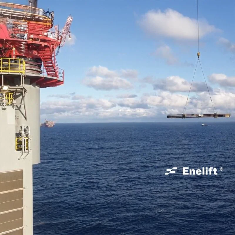 A large offshore oil platform at sea with a crane lowering a long cylindrical object, possibly a pipe or equipment, into the water. The sky is partly cloudy.