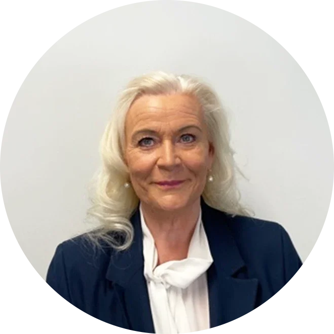 An older woman with long white hair, wearing a dark blazer and a white blouse, standing against a plain white background.