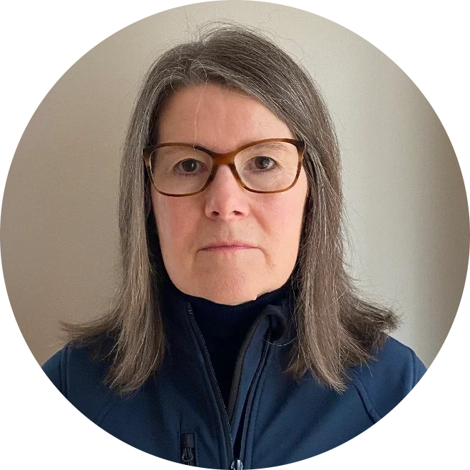 A woman with shoulder-length gray hair and glasses, wearing a black turtleneck and a dark blue jacket, posing against a plain light-colored wall.