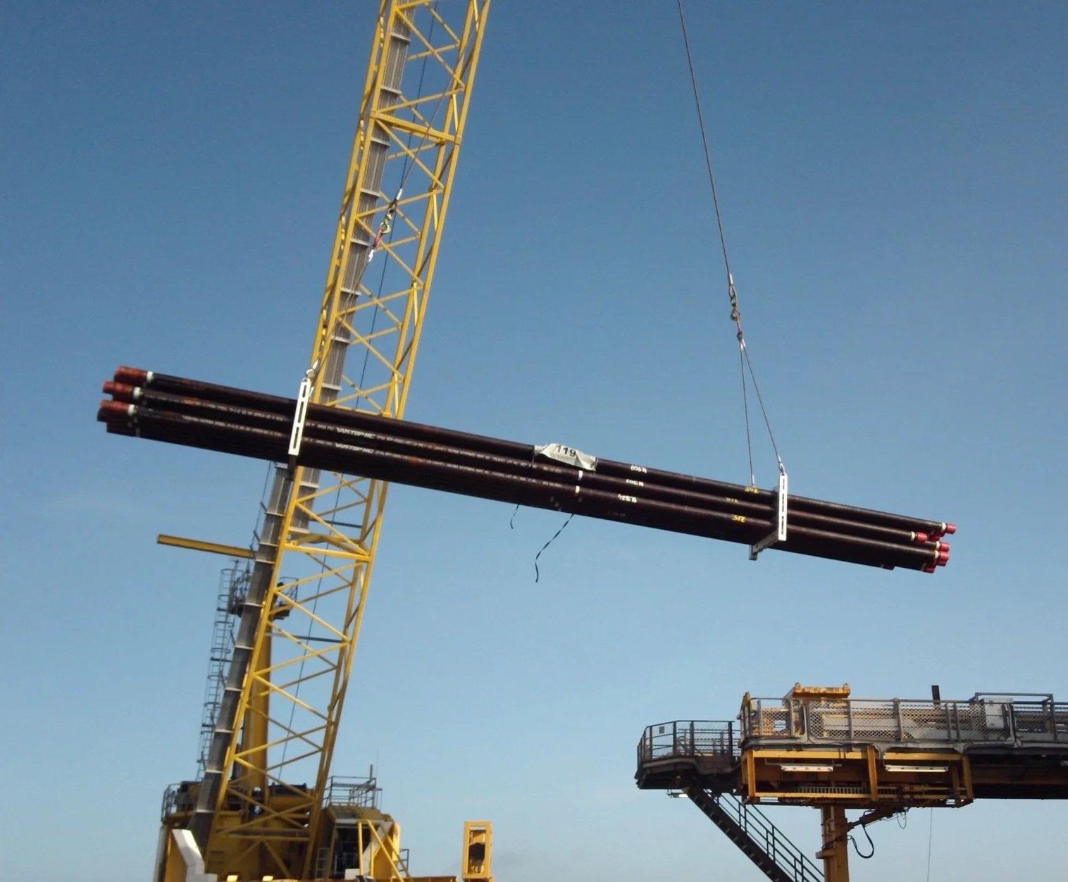 A construction crane lifting several long steel pipes against a clear blue sky.