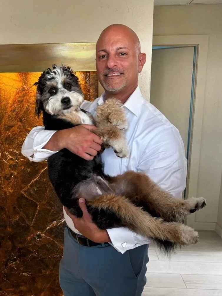 A man wearing a white shirt holding two small dogs in his arms. One dog is black and white, and the other is fuzzy and light-colored. The background features a brown marble wall and a cream-colored room.