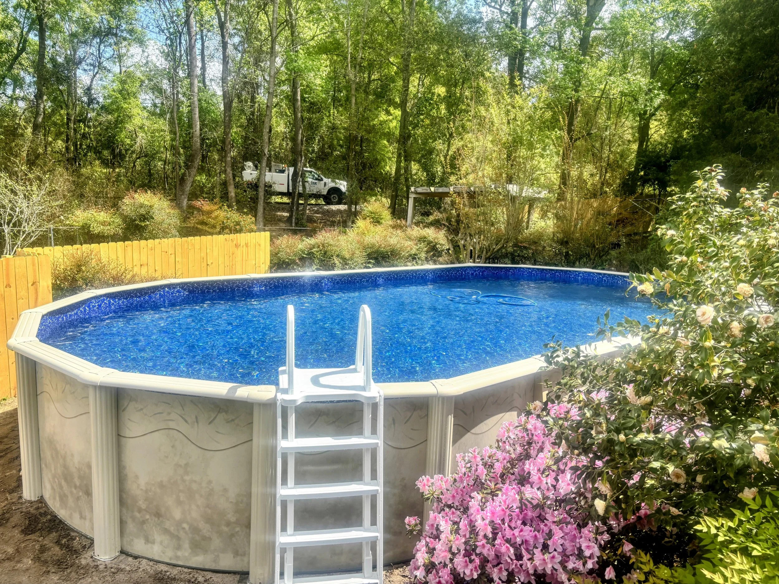 Above-ground swimming pool with white ladder, backyard flowers, and wooden privacy fence.