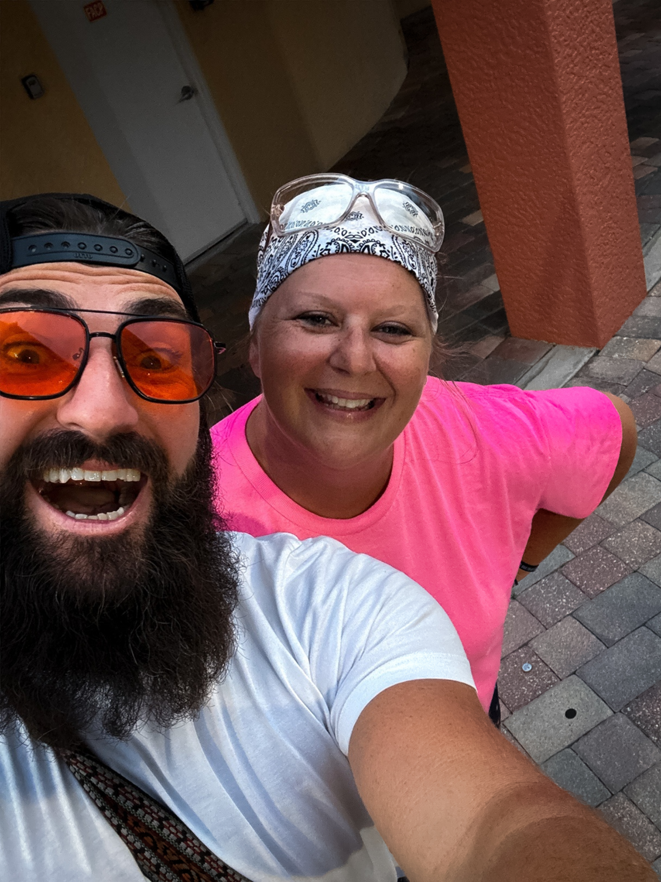 A man with a beard and mustache wearing orange-tinted sunglasses, a black cap worn backward, and a white shirt, smiling with an open mouth.