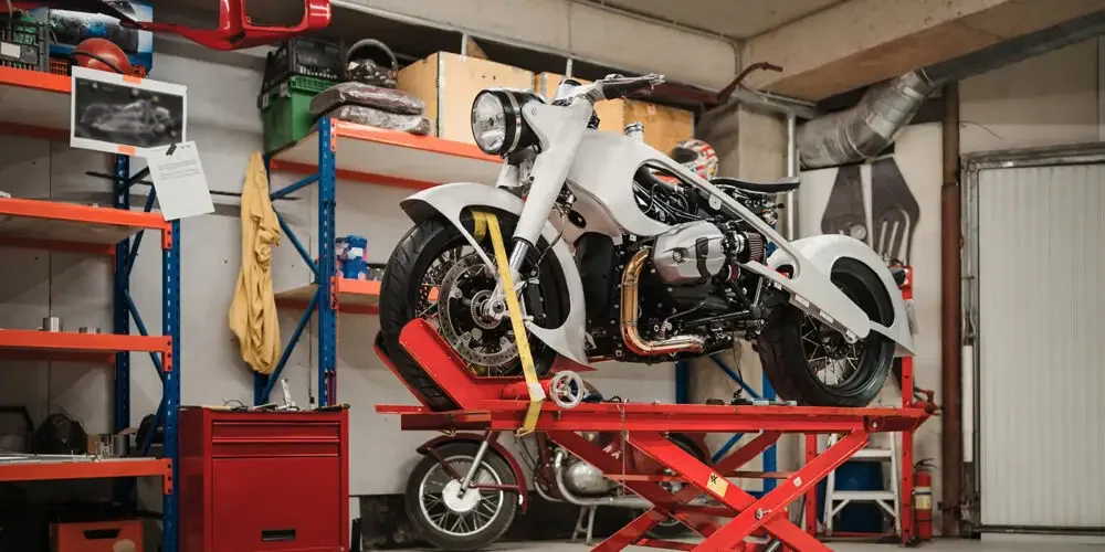 A white motorcycle on a red repair lift inside a garage or workshop with tools and shelves in the background.