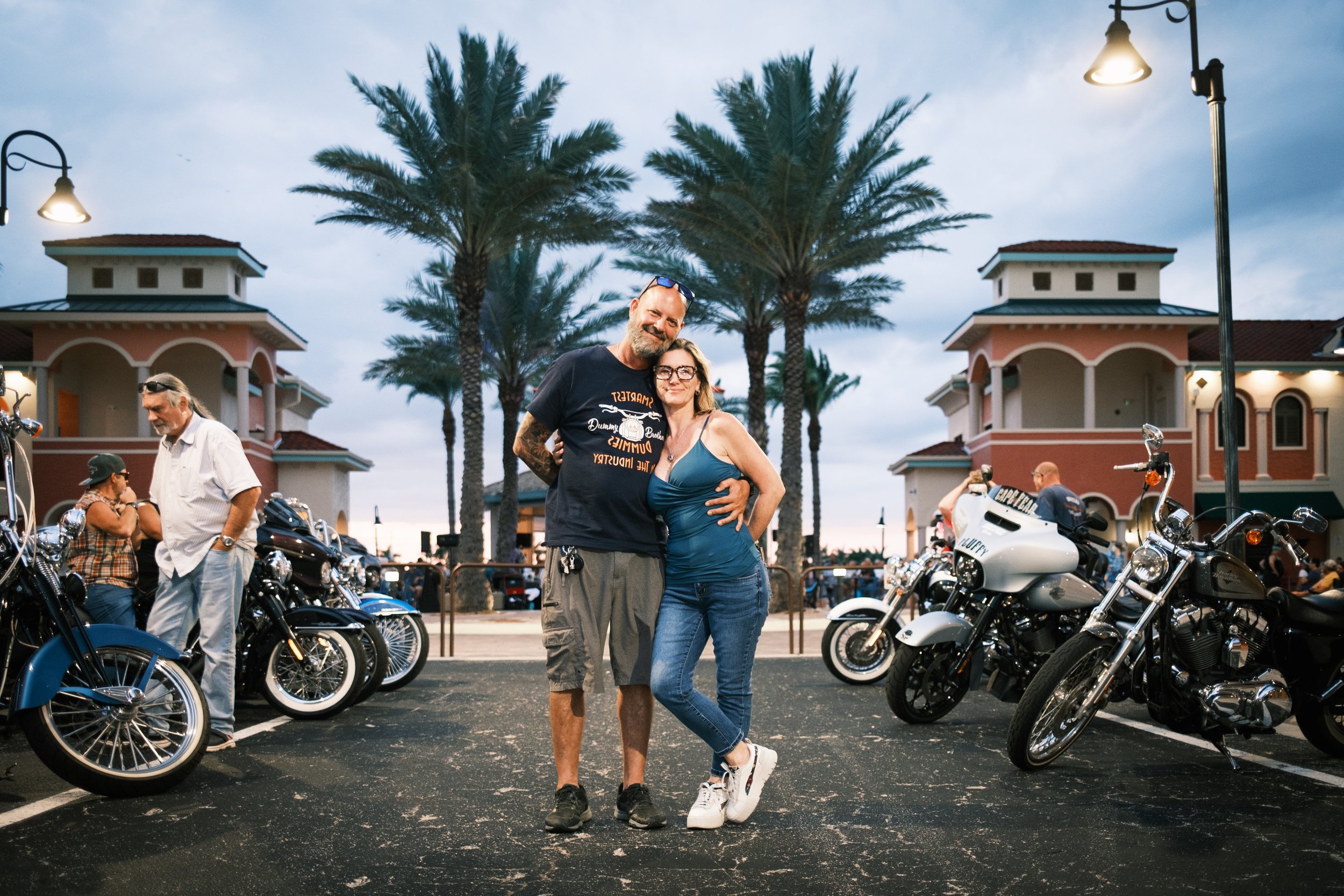 A couple stands close together in a parking lot filled with motorcycles, behind them palm trees, pink and beige buildings, and street lamps are visible under a partly cloudy sky.