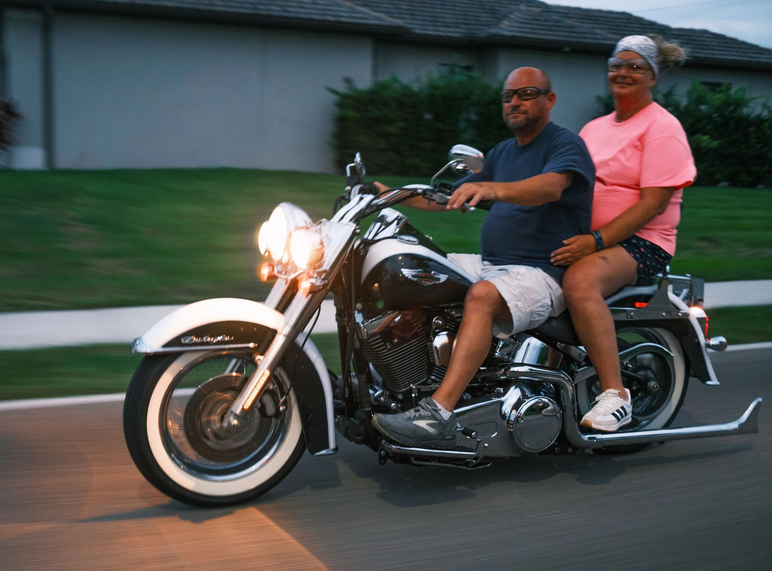 A man and a woman riding a black and white motorcycle together on a street during dusk, with the woman sitting behind the man, both smiling and looking forward.
