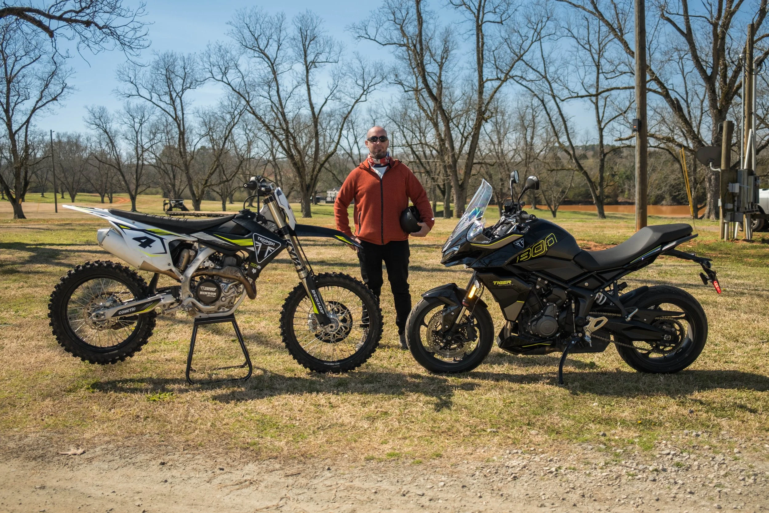 A man standing outdoors between two motorcycles in a park with leafless trees and a blue sky.