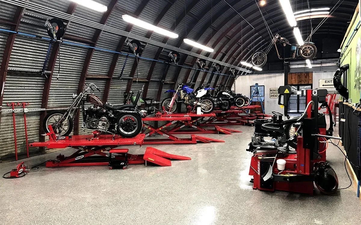 An indoor motorcycle repair shop with several motorcycles on red hydraulic lifts and a motorcycle wheel balancing machine in the foreground. The shop has a curved metal ceiling with fluorescent lighting and a corrugated metal wall with hanging bike helmets.