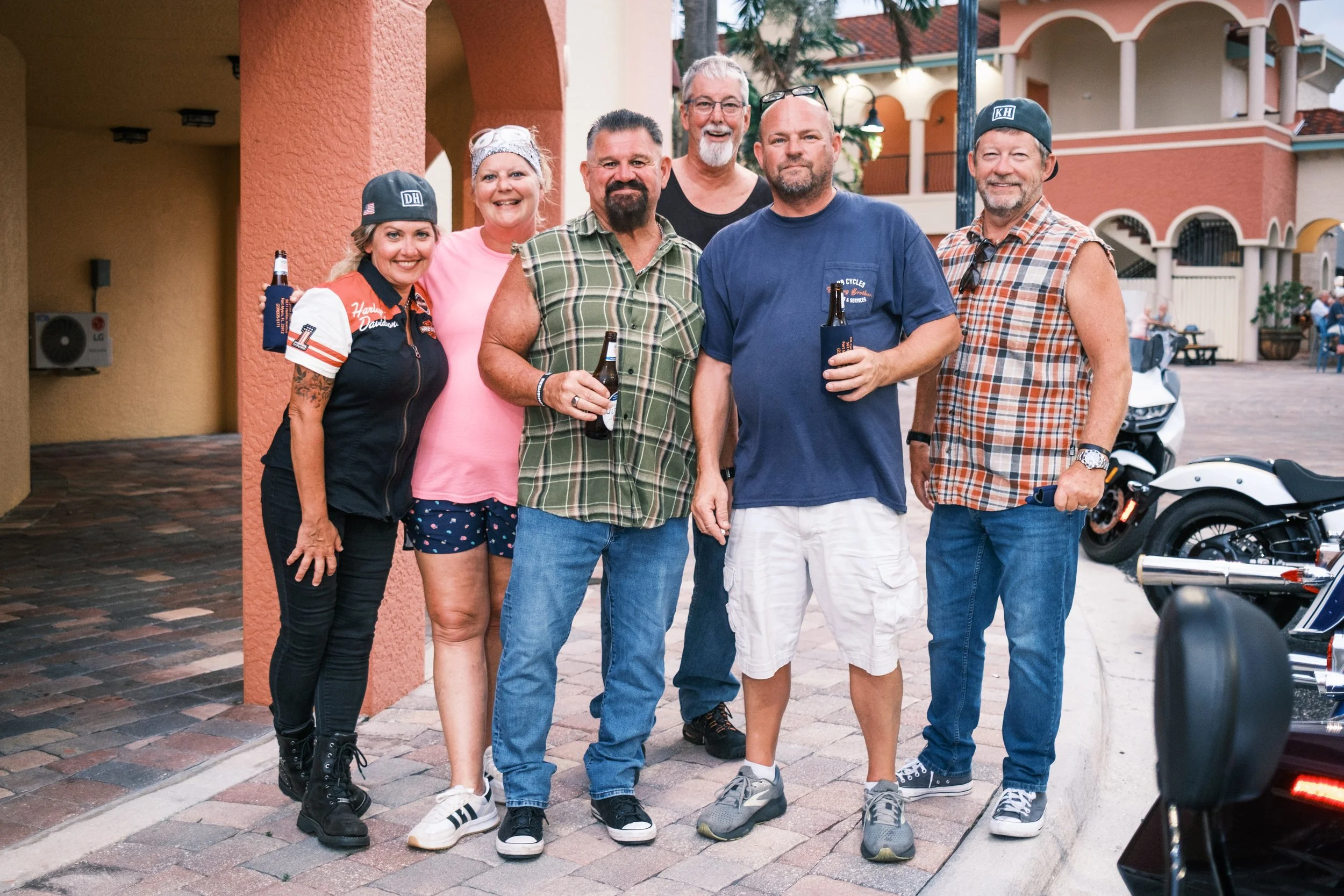 Group of seven diverse adults standing together outdoors with buildings in the background, smiling and holding drinks.
