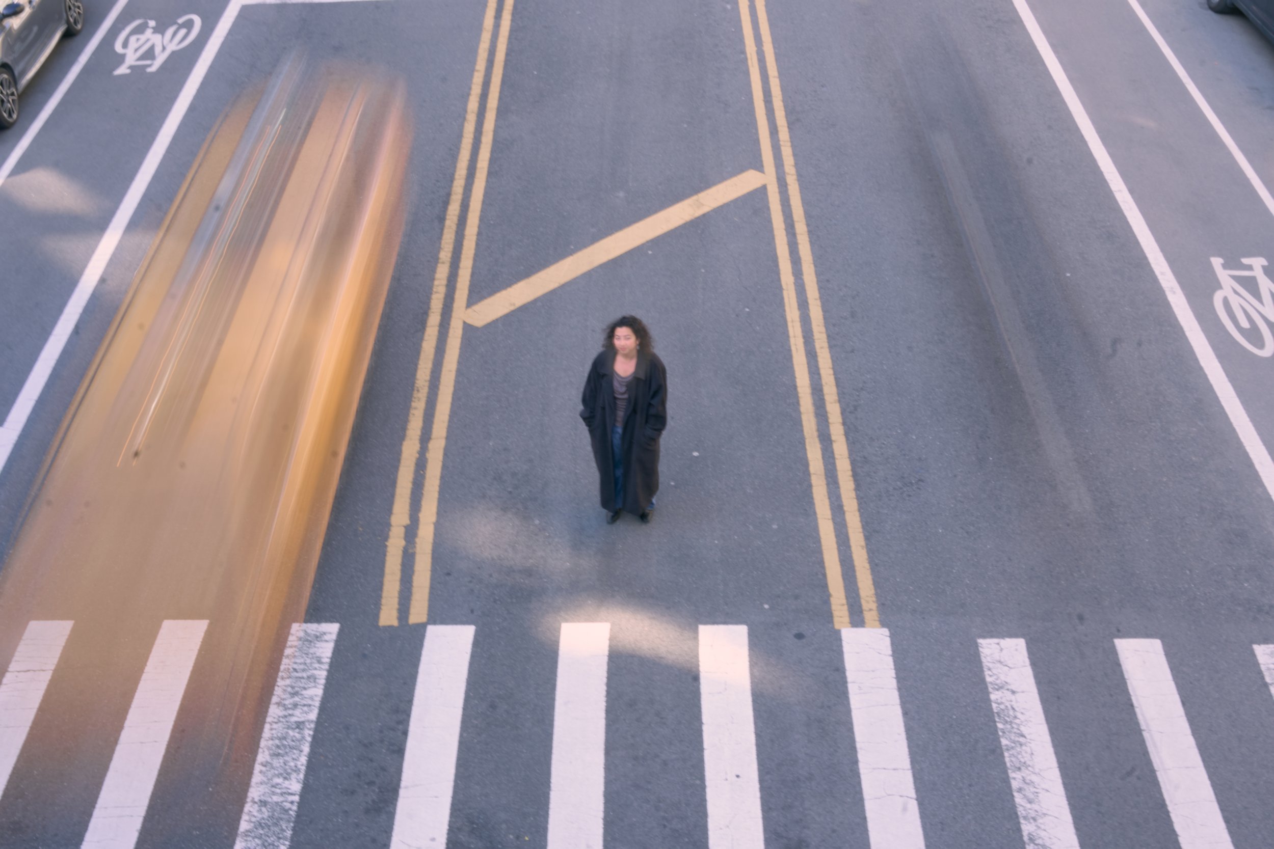 A woman standing in a parking lot near a designated parking space for bicycles, with some cars parked nearby. The photo is taken from above, with motion blur visible on a moving vehicle on the left side.