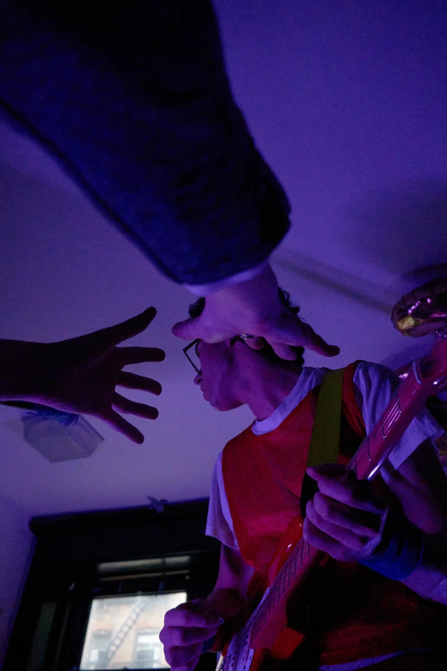 A person playing an electric guitar, wearing a red and white shirt, with a yellow strap, in a dimly lit room with purple lighting. The person's face is turned to the right, and they are surrounded by another person with extended fingers reaching towa
