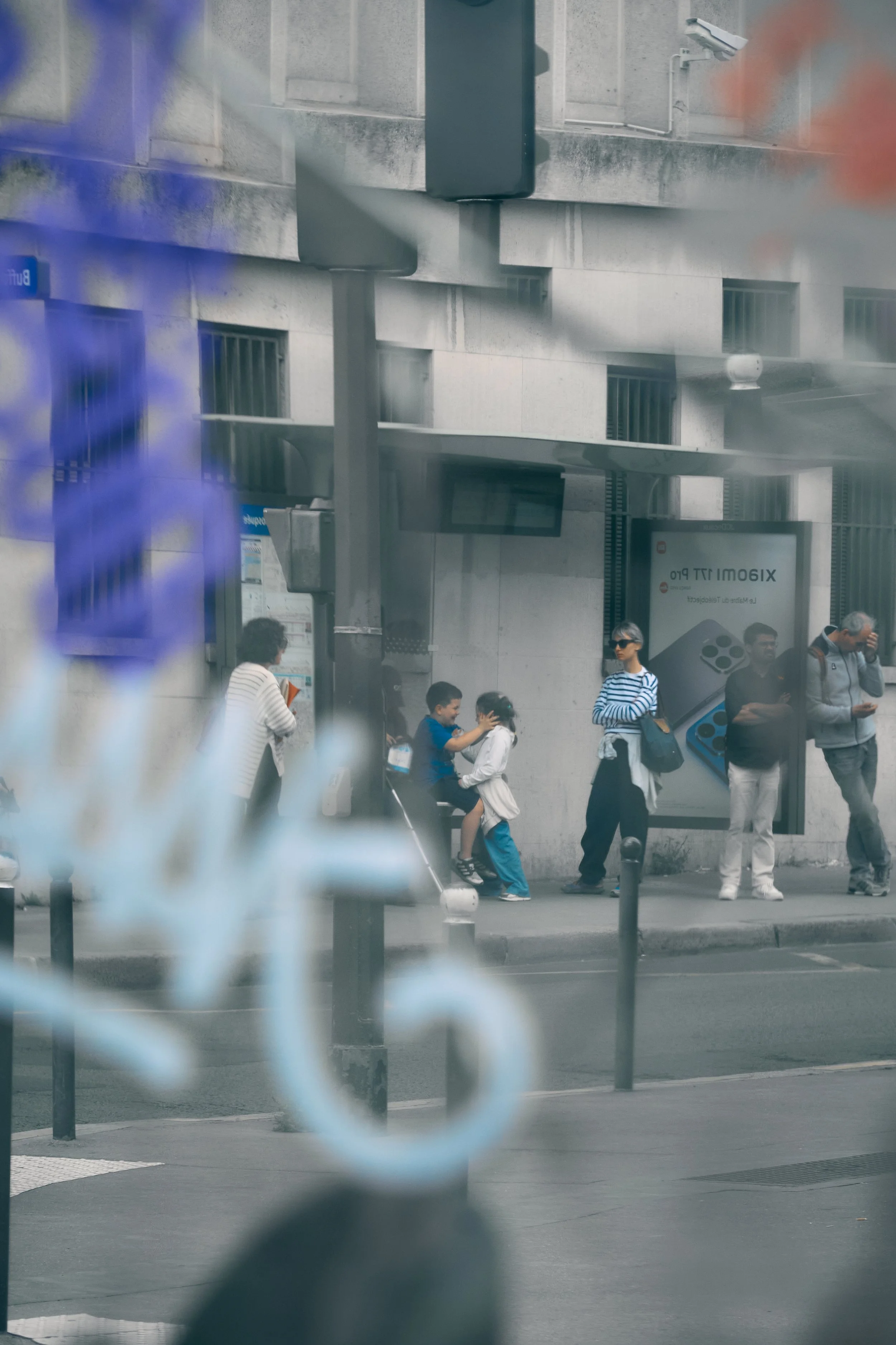 People waiting at a bus stop in front of a stone building, with a sign advertising a product, and a security camera on the building.