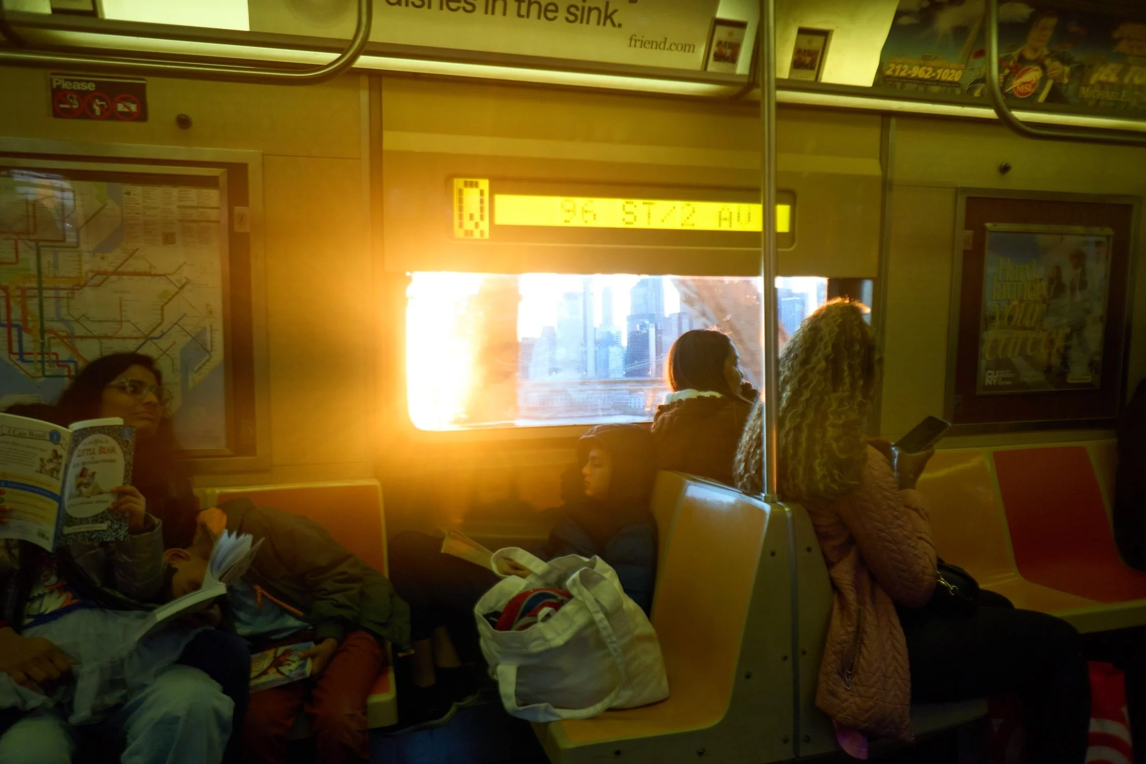People sitting in a subway car, some reading books and others using smartphones, with a city skyline visible through the window.