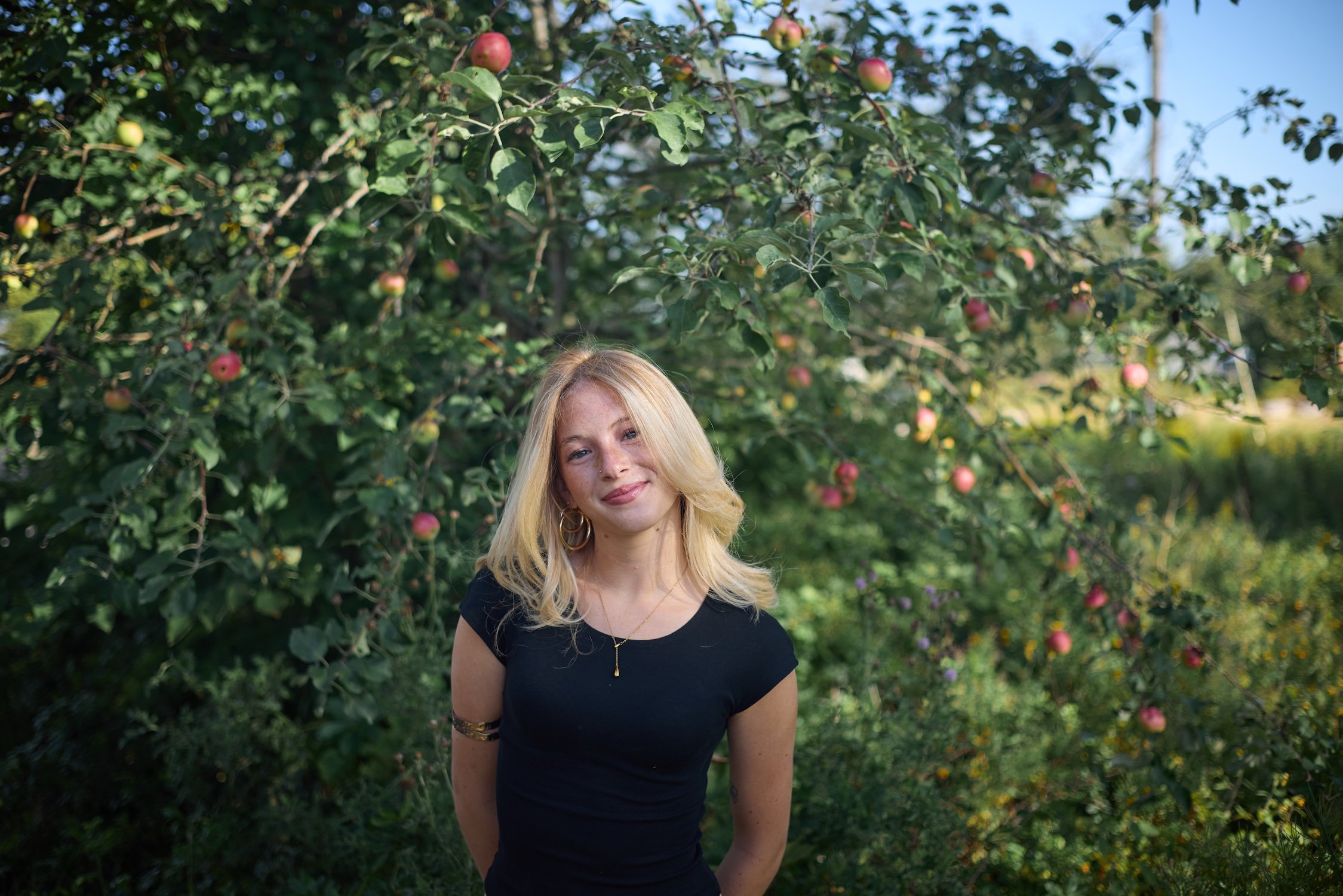 A young woman with blonde hair and hoop earrings standing outdoors in front of an apple tree with ripe apples.