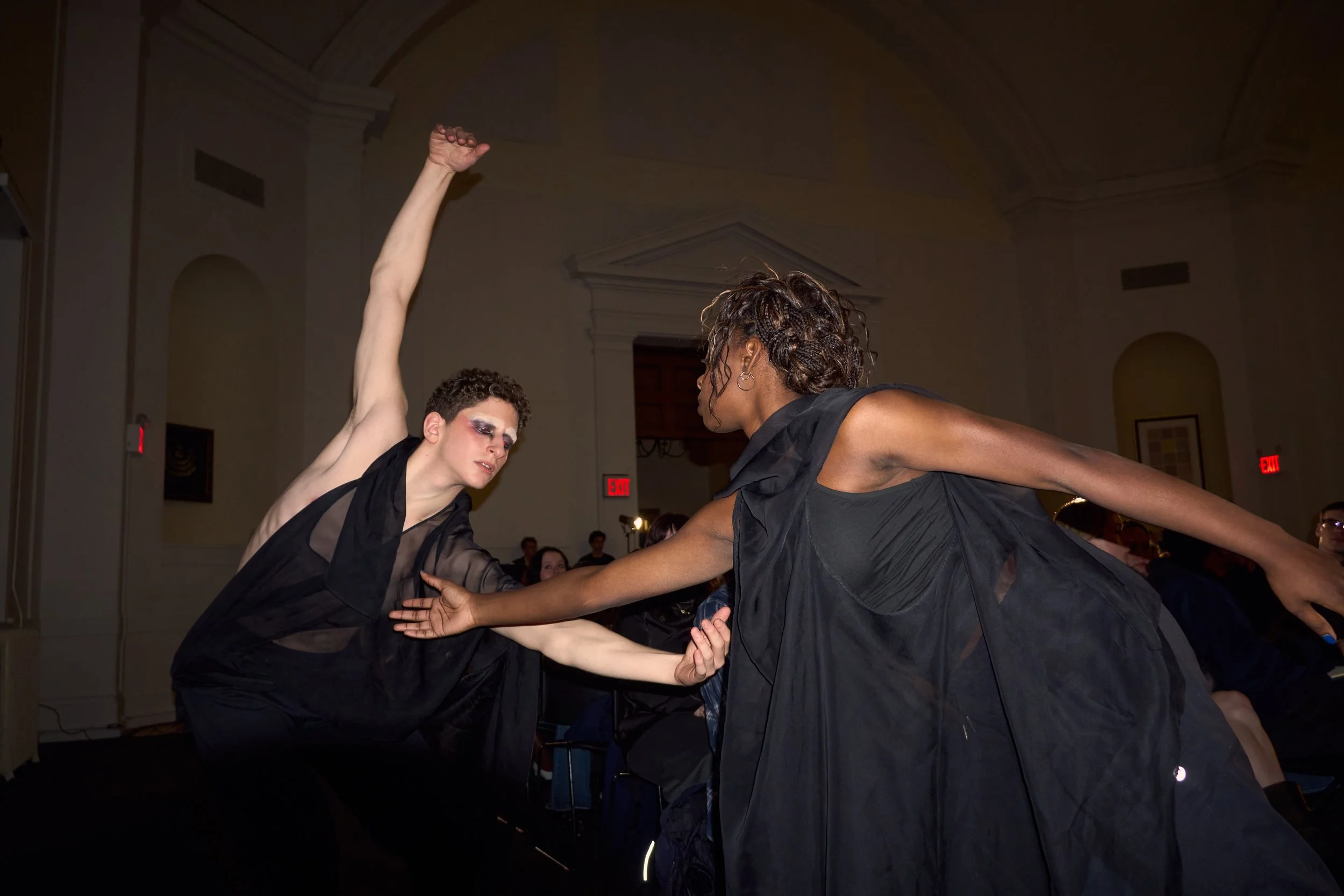 Two dancers in black outfits performing a dance in a grand indoor setting, with audience members watching in the background.