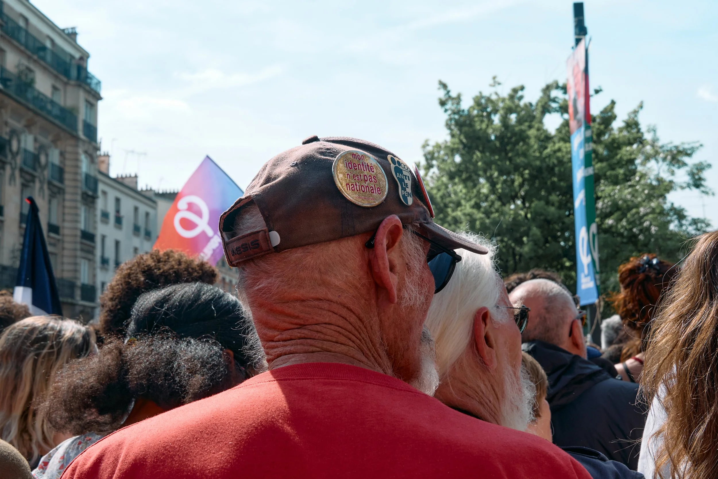 Crowd of people at an outdoor event with a man wearing a brown baseball cap with campaign stickers, including one that says 'mon identité n'est pas nationale,' in front of buildings and trees during daytime.