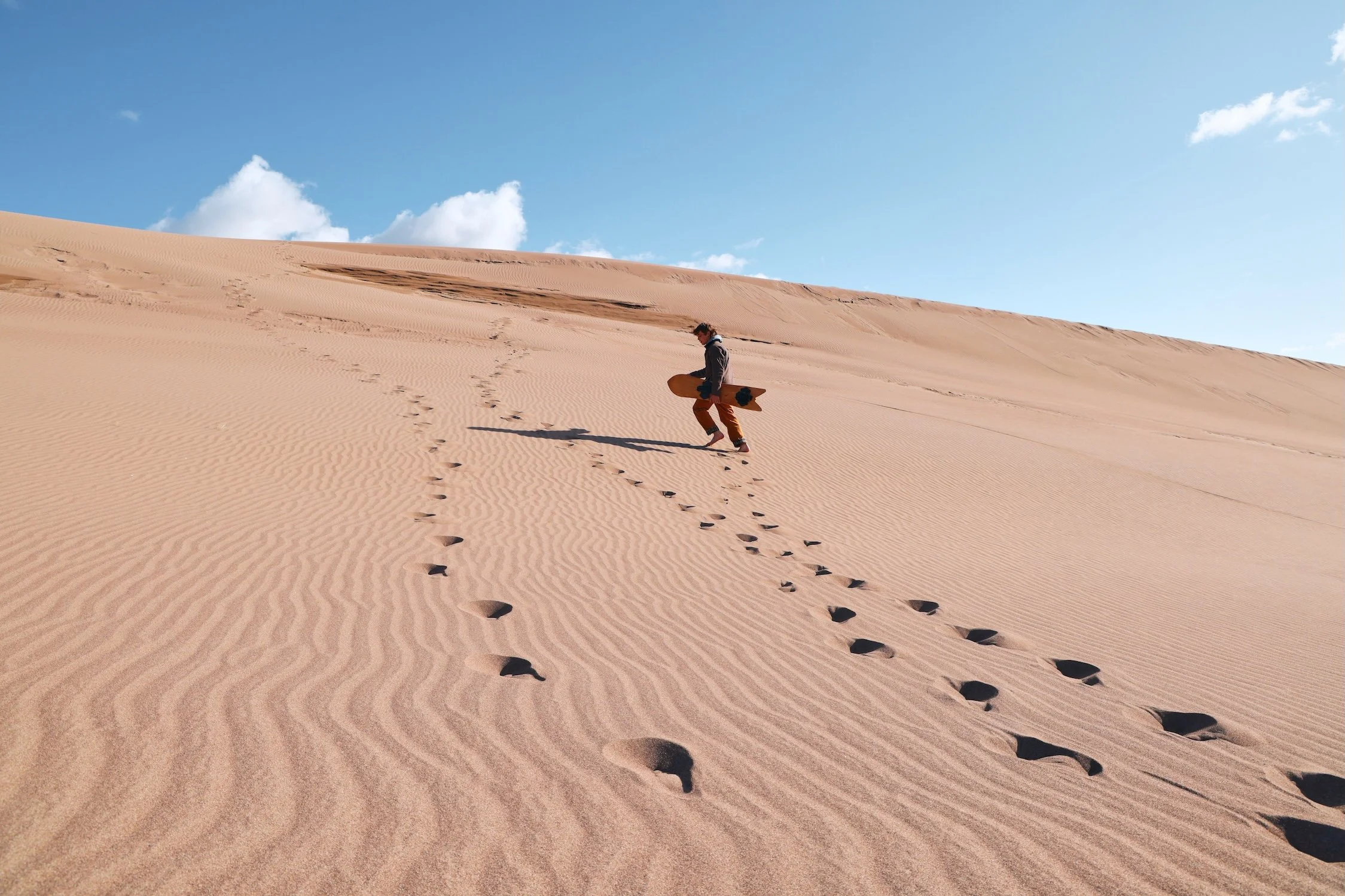 A person walking on sand dunes holding a skateboard, leaving footprints in the sand, under a blue sky with scattered clouds.