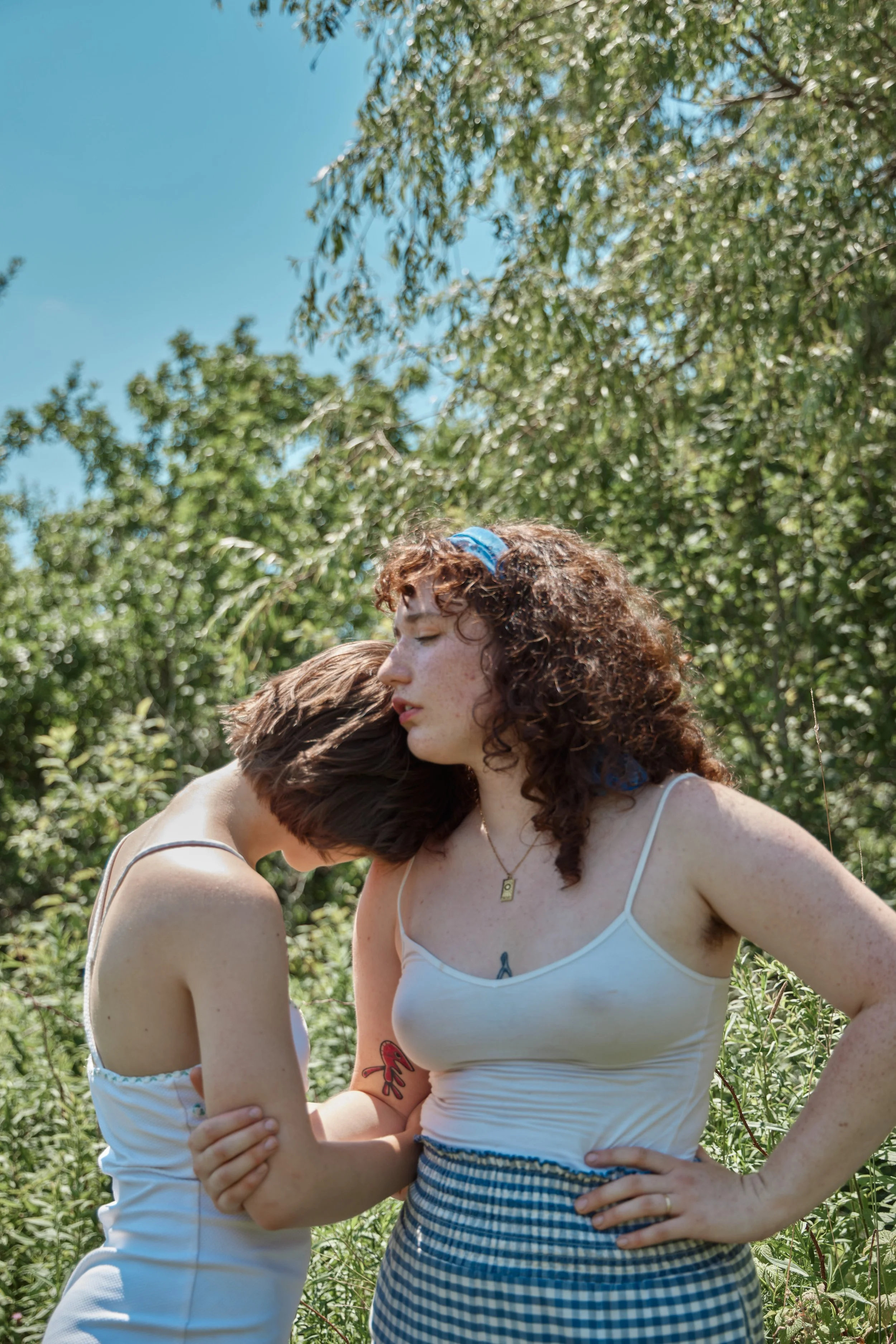 Two young women with curly hair embracing outdoors among greenery, one with a blue headband and the other with a tattoo on her arm, wearing white tank tops.