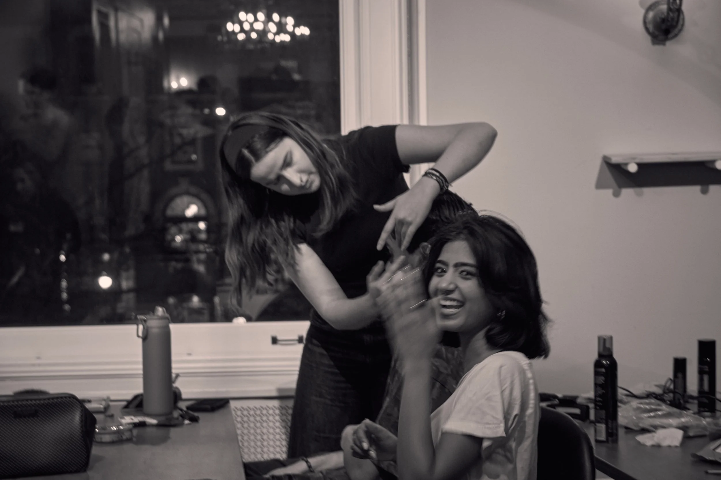 Two young women, one with long wavy hair and dark eyebrows, are sitting at a table, smiling and looking at the camera. The woman with wavy hair is holding a drink, and the other woman is leaning in, with her head resting on the shoulder of the woman 
