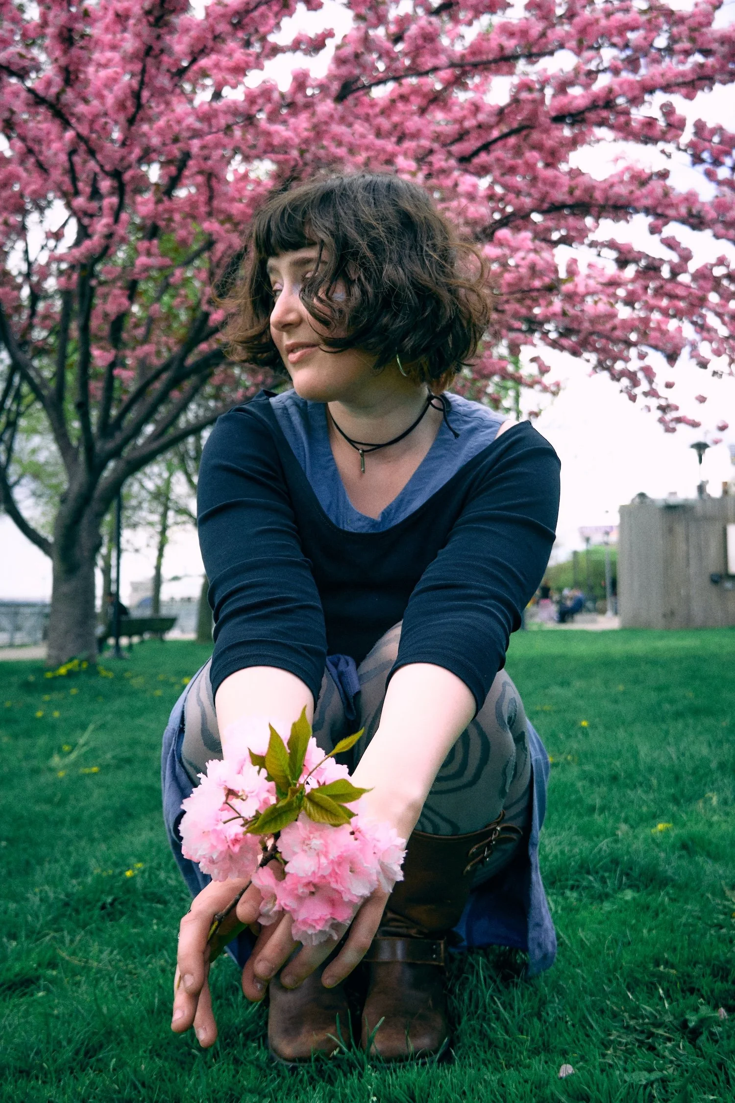 A woman with curly brown hair in outdoor park during spring, kneeling on green grass, holding pink cherry blossoms, with a large pink cherry blossom tree in the background.