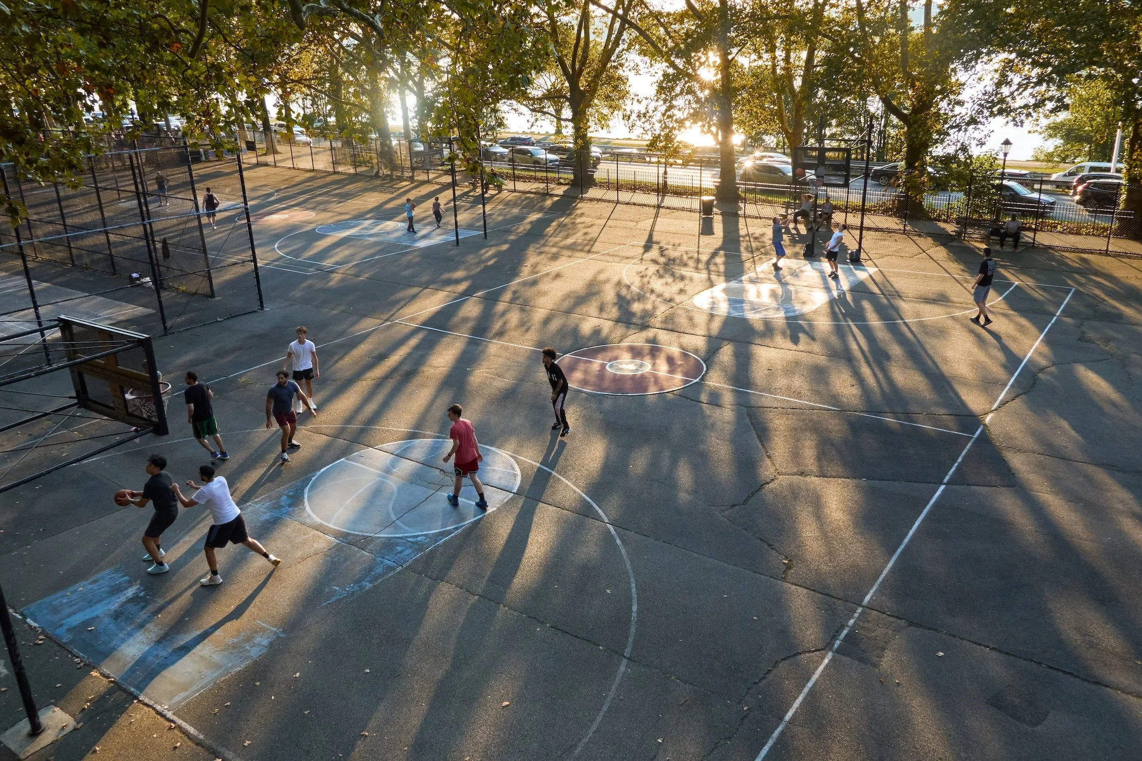 Young people playing basketball on outdoor courts with trees and parking lot in the background during sunset.