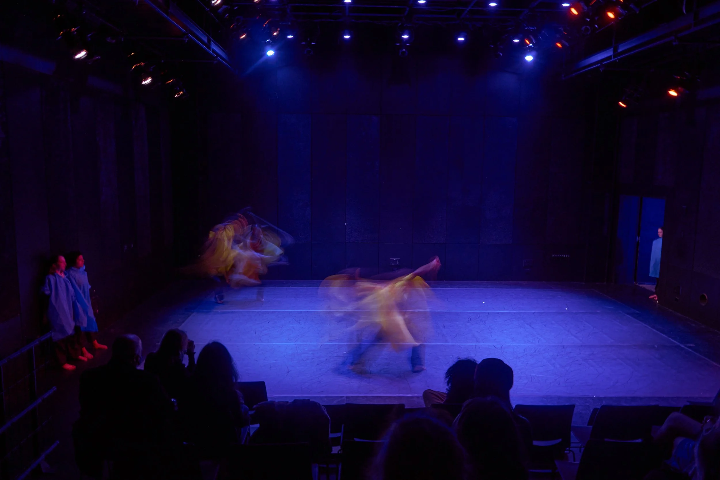 Stage with blue and purple lighting, audience seated in front, and dancers performing with blurred motion, indicating movement.