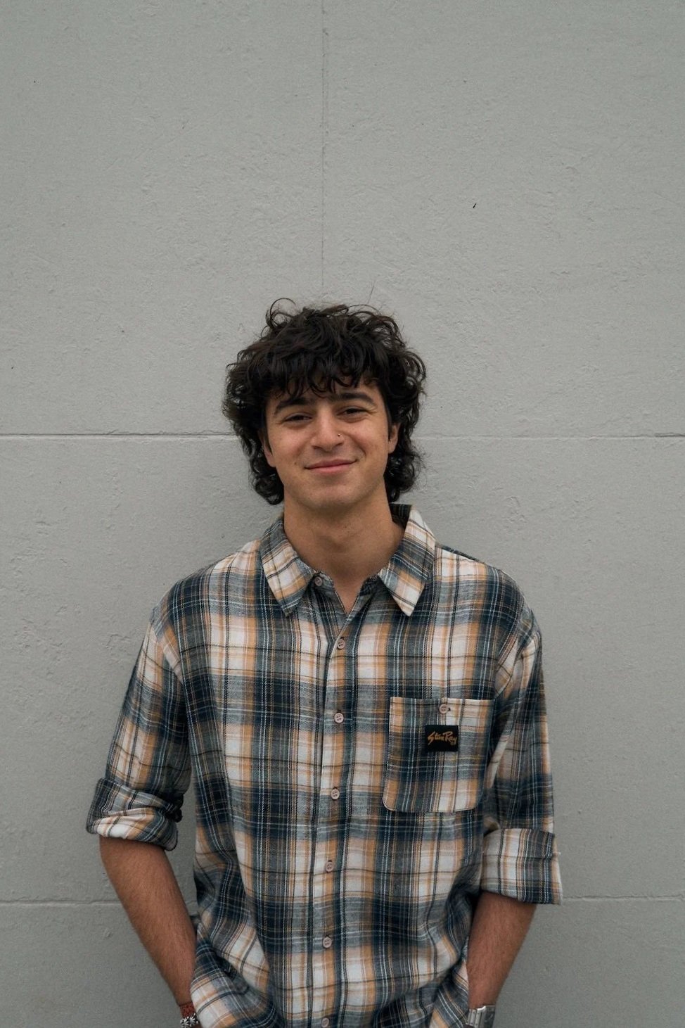 Portrait of a young man with curly dark hair smiling, standing against a plain light gray wall, wearing a plaid button-up shirt with beige, blue, and white colors.
