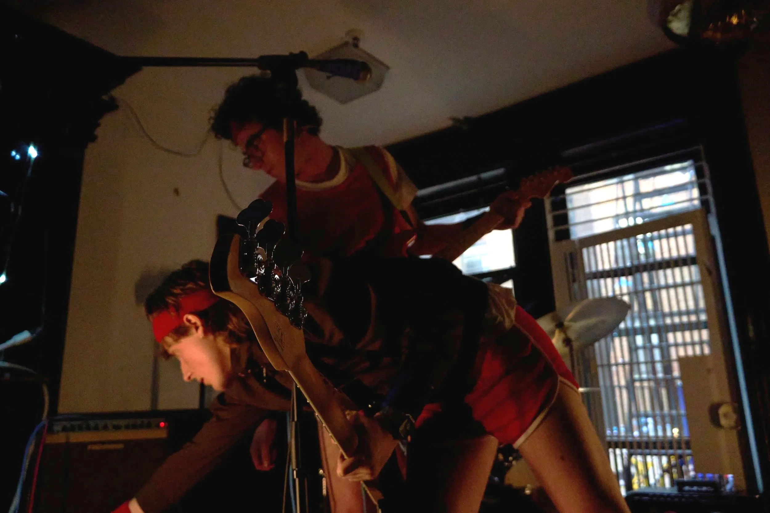 A person with curly hair and glasses playing an acoustic guitar in a dimly lit room, with a window and blinds in the background.
