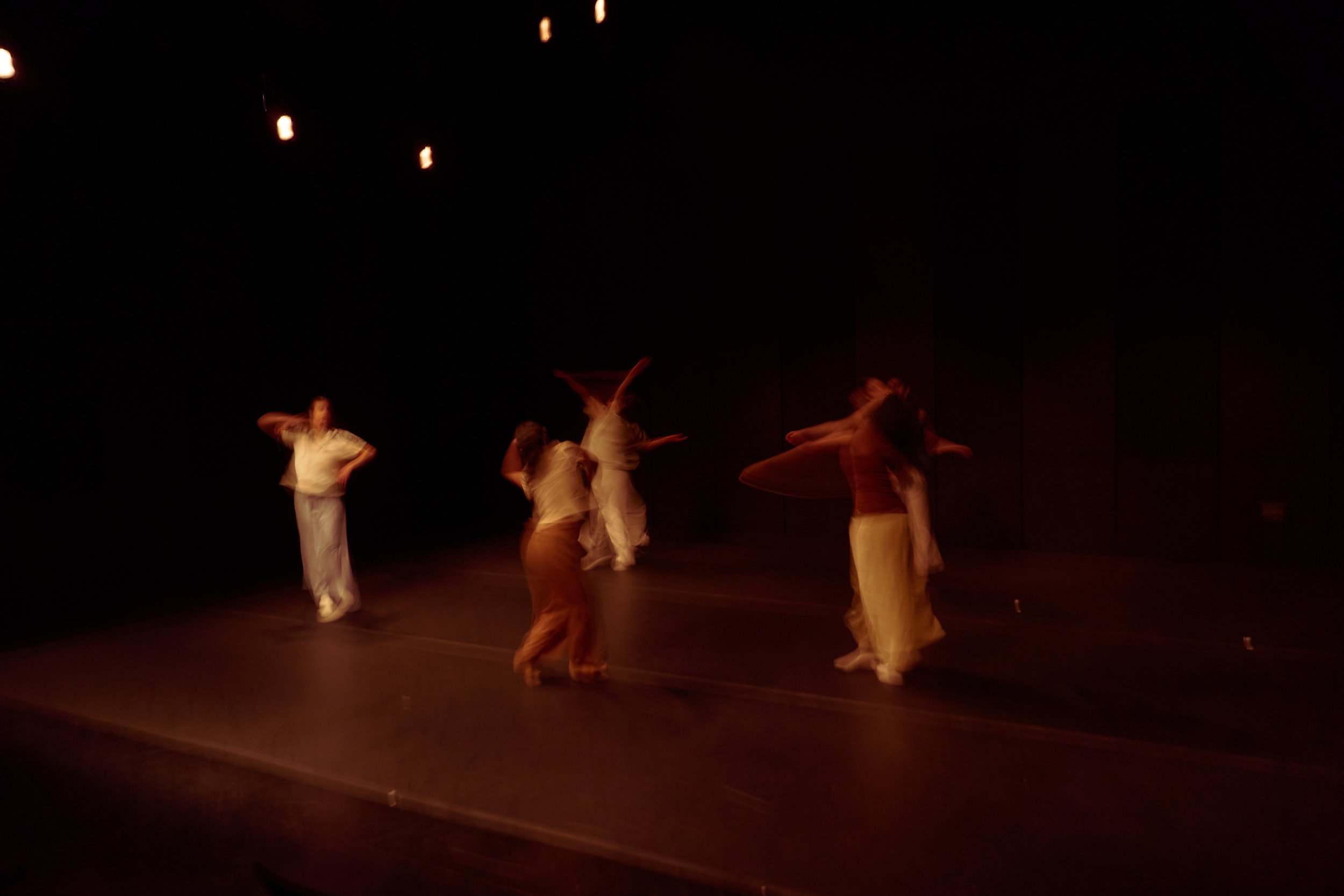 A group of dancers performing on stage in dim lighting, wearing light-colored costumes, with some dancers in motion and others standing still. The stage appears dark with minimal lighting focus.