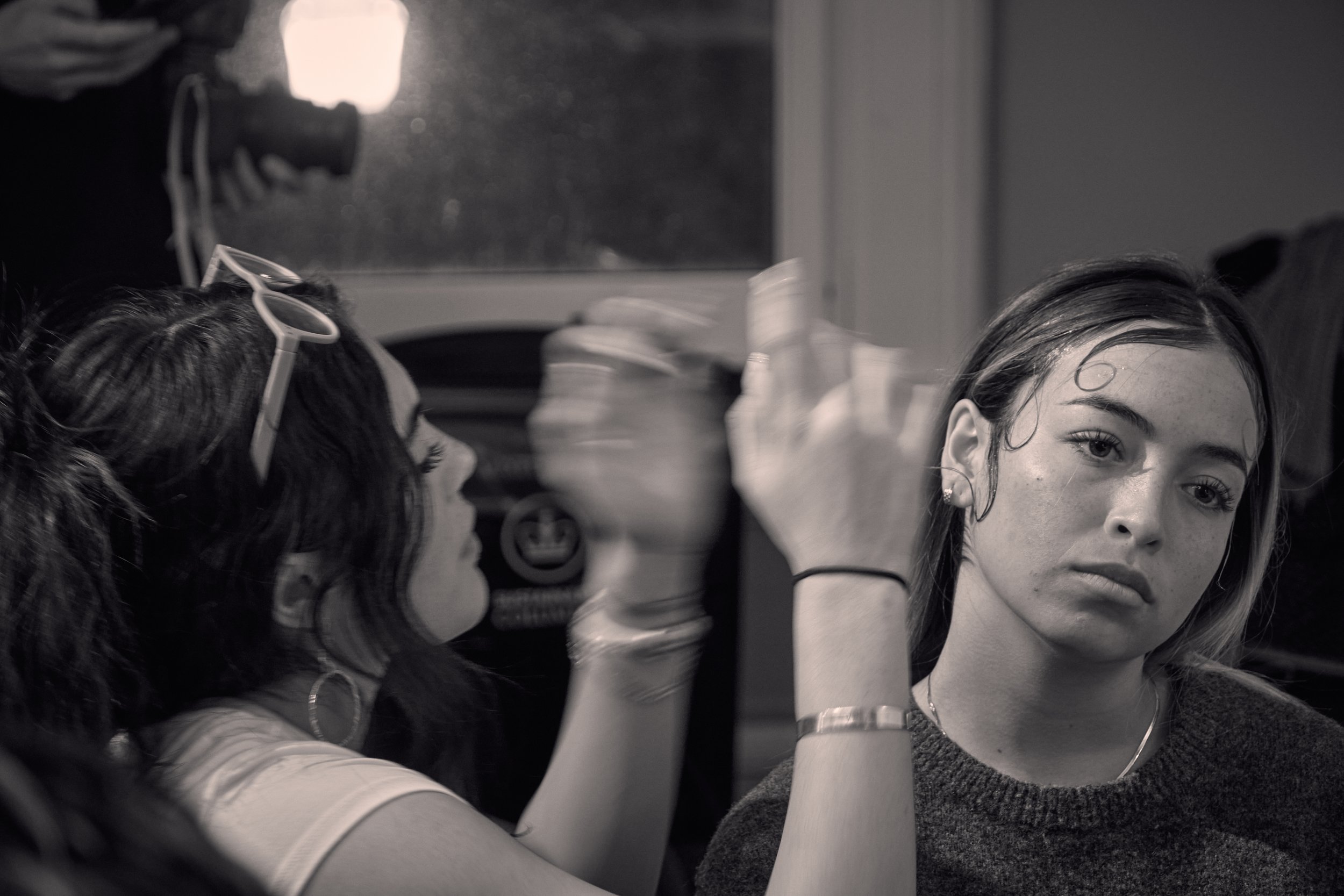 A woman with wet hair looking into a mirror, applying makeup or skincare. The image is in black and white, with a blurred background.