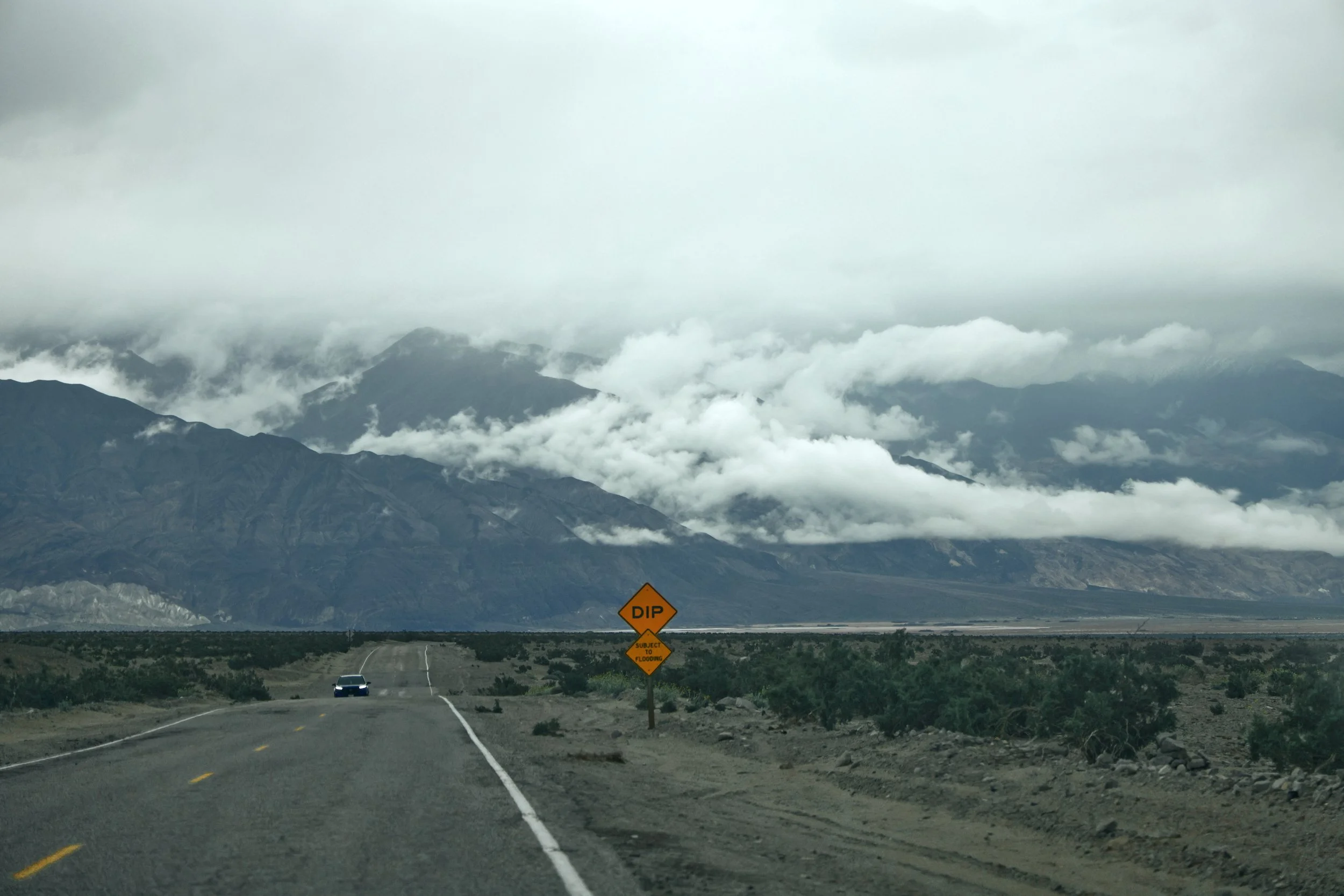 A long two-lane highway running through a desert landscape with mountains in the background. Clouds cover the mountain peaks. On the right side of the road, there is an orange warning sign that reads 'DIP' and 'SUBJECT TO FLOODING'. A car is approach