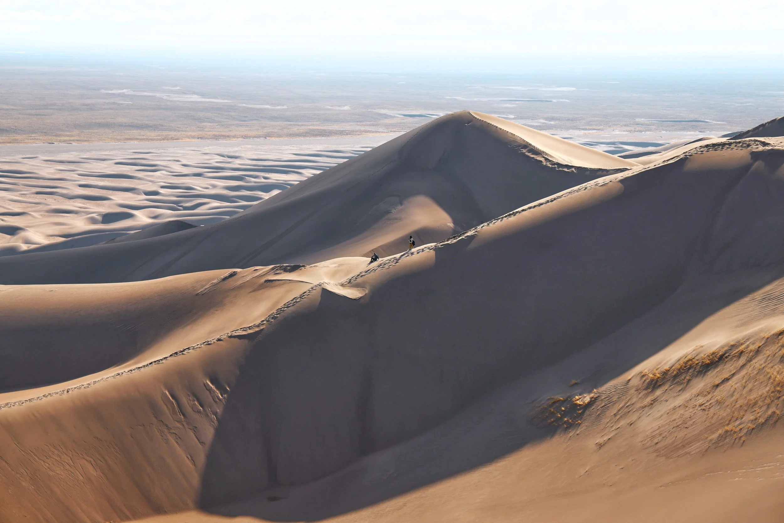 Sand dunes in a desert landscape, with a person standing near the peak of a dune and footprints in the sand.