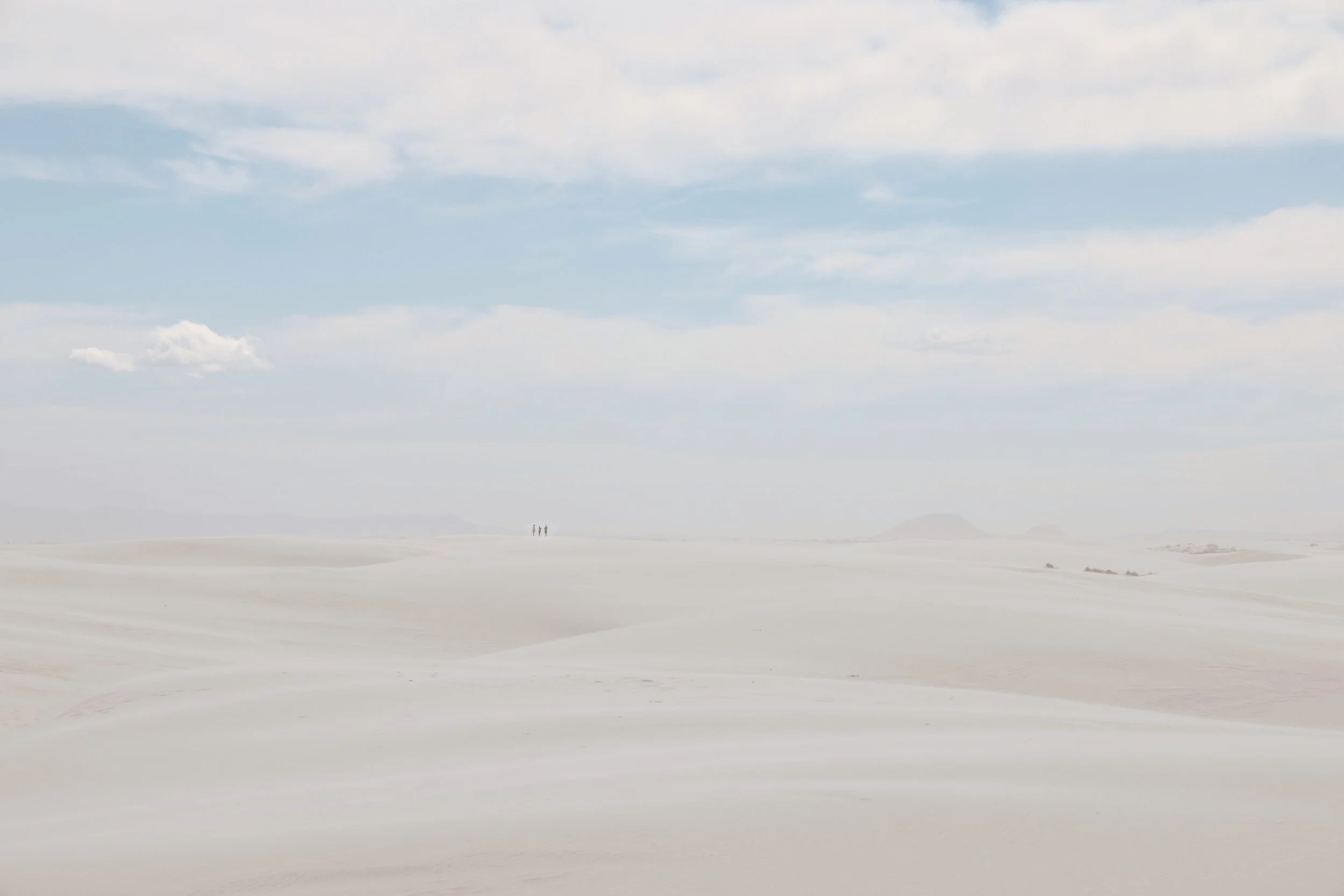 Wide landscape of white salt flats under a blue sky with scattered clouds, with two distant figures walking across the salt flats.