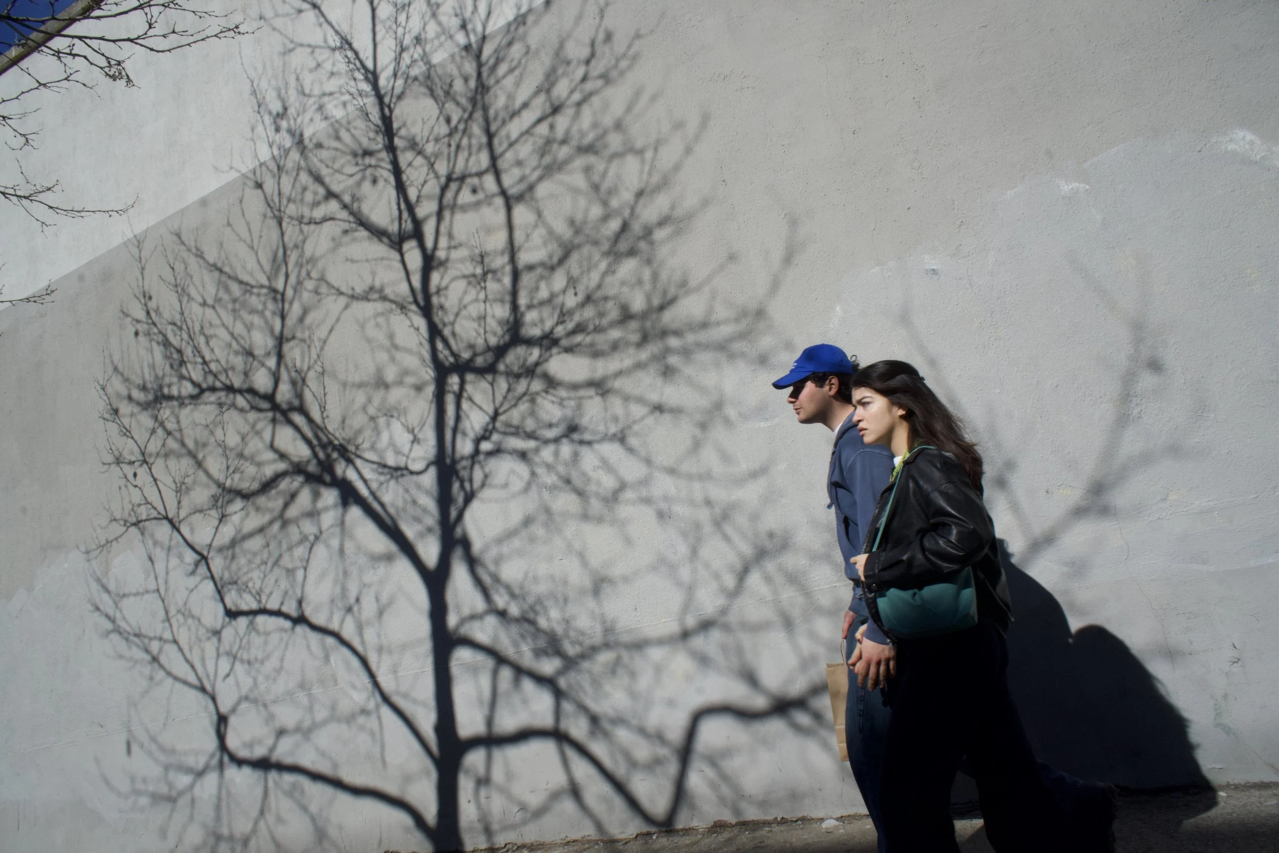 Two people, a man in a blue cap and a woman, walking side by side against a white wall with a leafless tree casting shadows on it.