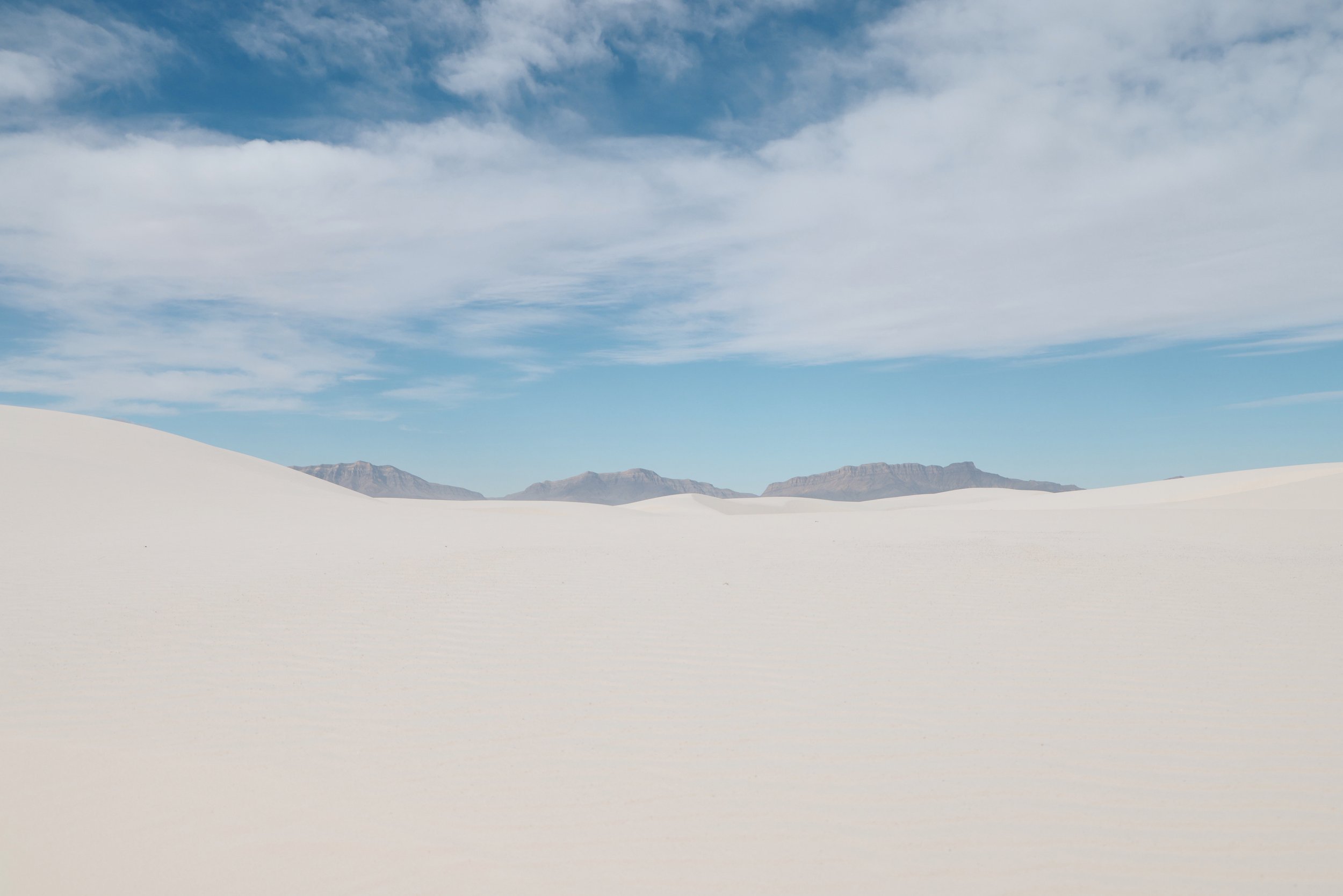 A desert scene with white sand dunes, distant mountains, and a partly cloudy sky.