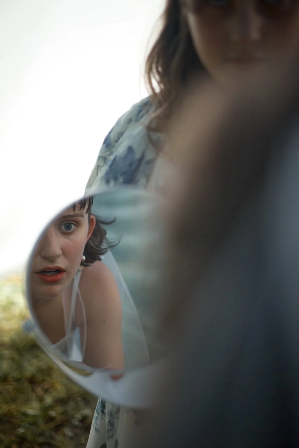 A woman and a girl looking into a reflective surface, possibly a mirror, outdoors during daytime. The woman appears to be examining her reflection, while the girl looks directly into the mirror with a surprised or intrigued expression.