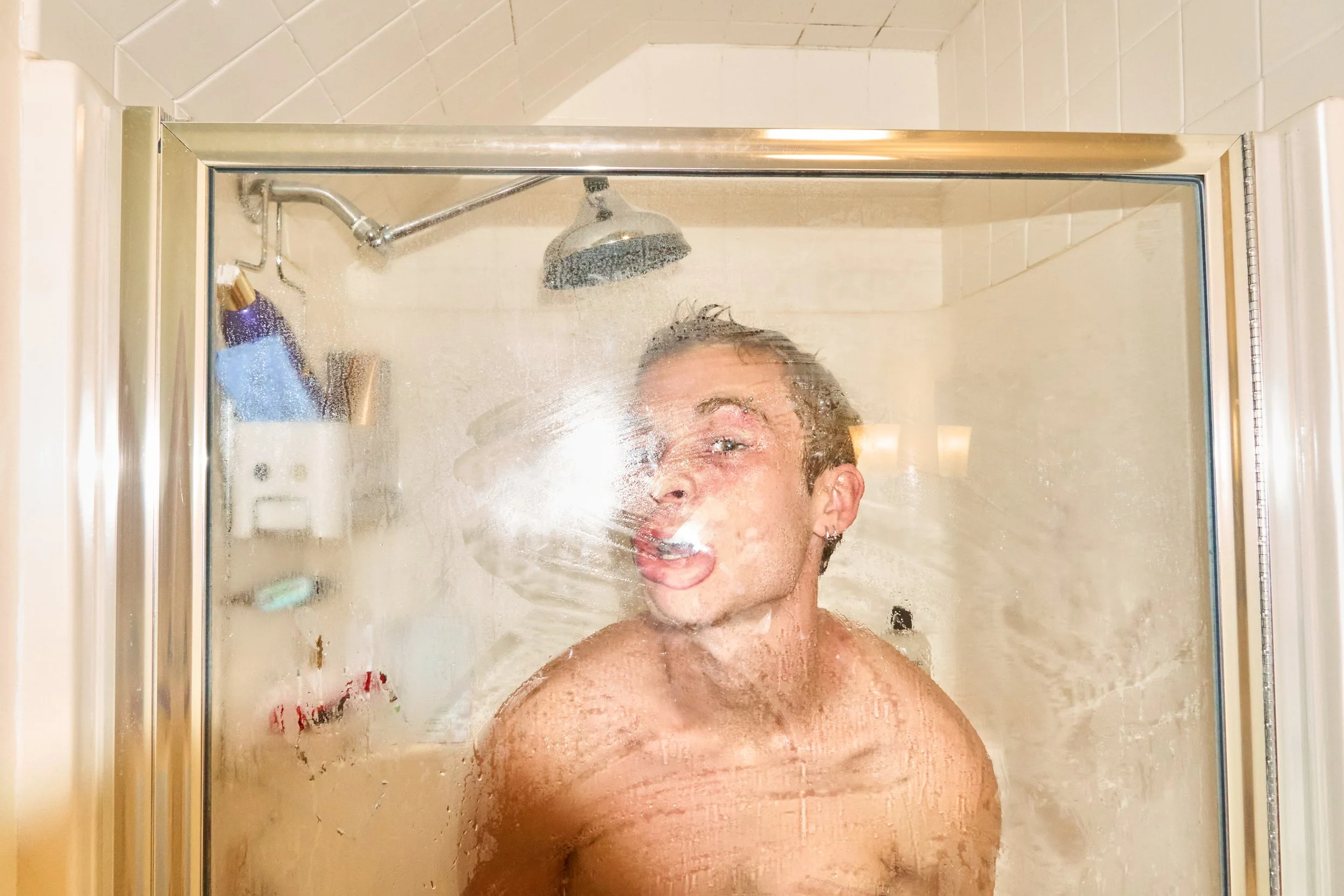 A person taking a shower with their head tilted back and mouth open, water running down their face, viewed through a glass shower door.