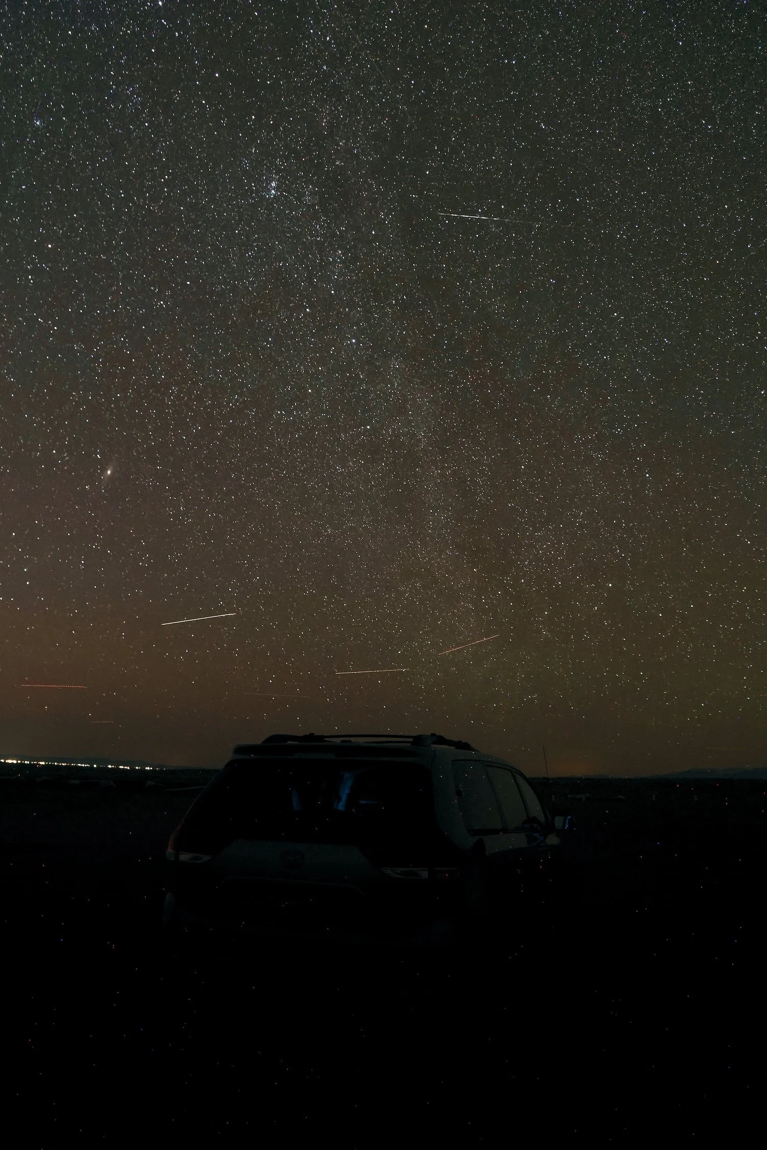 A night sky filled with numerous stars and a few shooting stars or meteors. The silhouettes of a car and a landscape are visible at the bottom.