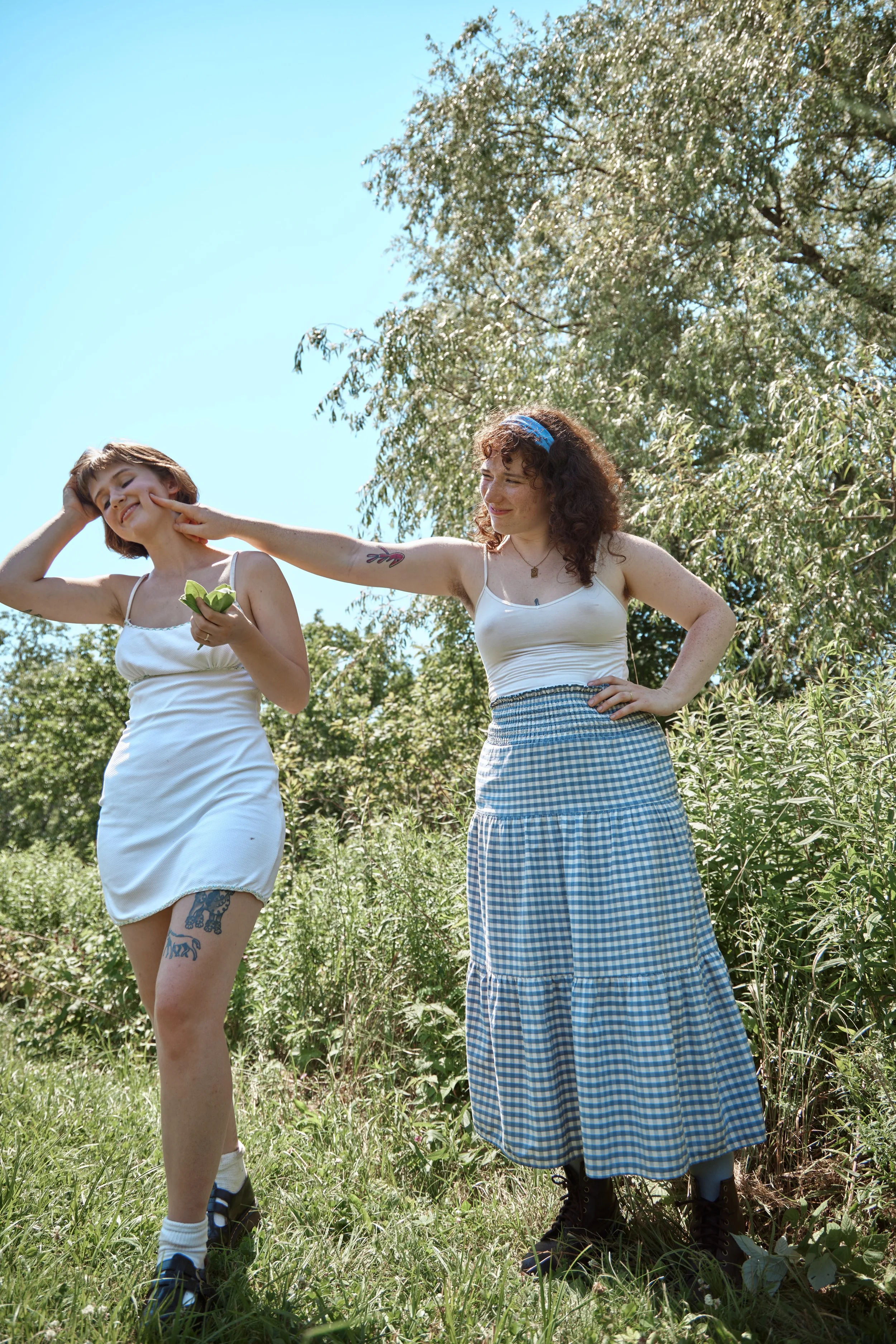 Two women standing outdoors in a grassy field surrounded by trees on a sunny day. One woman touches the other's face in a playful manner, both smiling. The woman on the left wears a white dress with tattoos on her leg, and the woman on the right wear