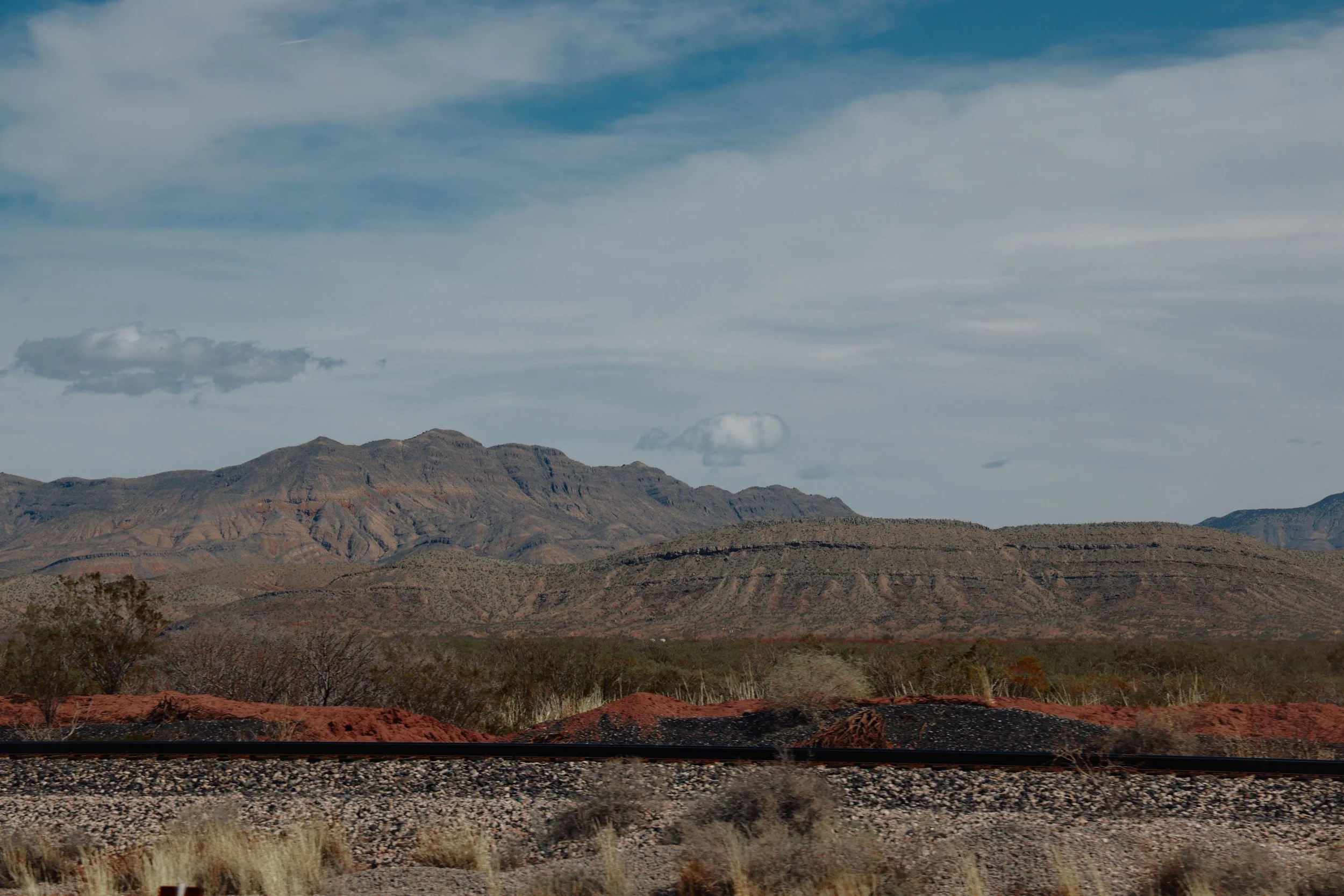 A desert landscape with reddish-brown mountains and sparse vegetation, under a partly cloudy sky.