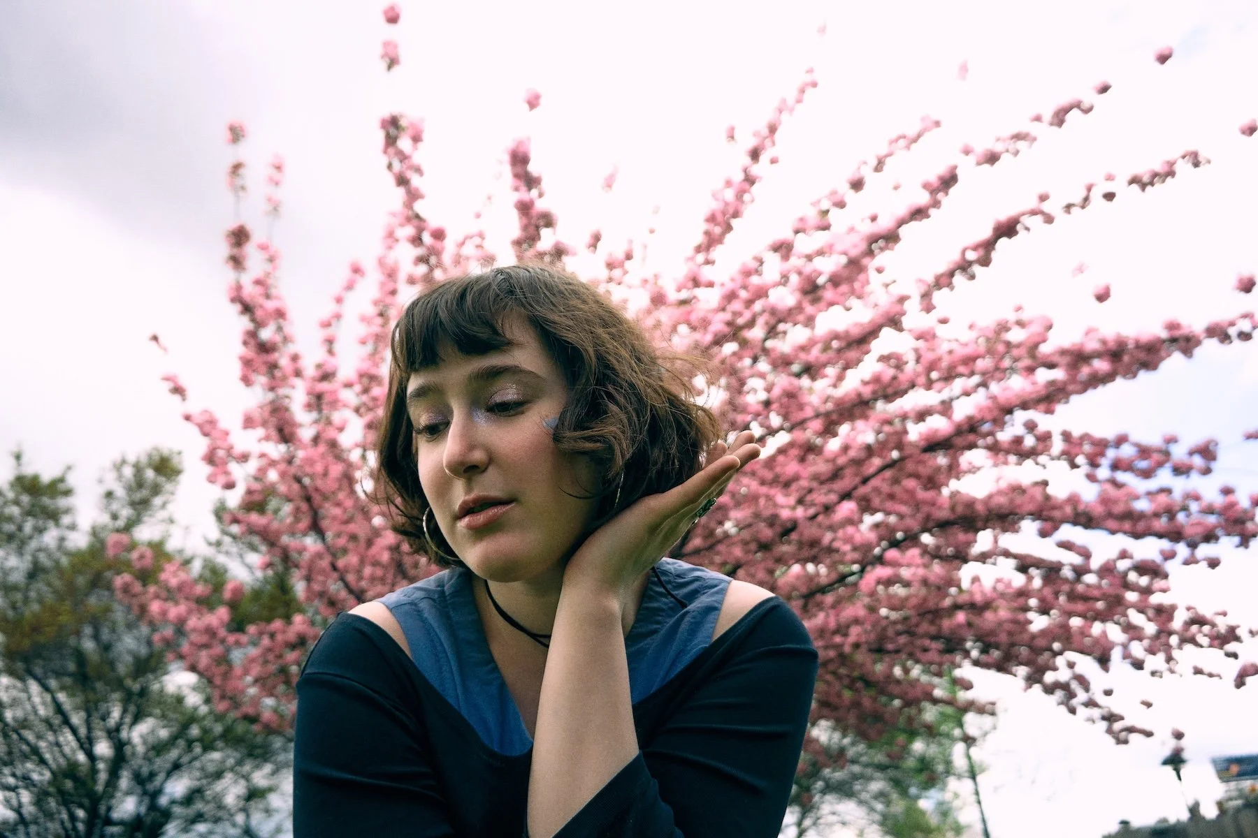 A young woman with short curly hair, wearing a dark top and a choker, posing pensively in front of blooming pink cherry blossom trees in an outdoor park.