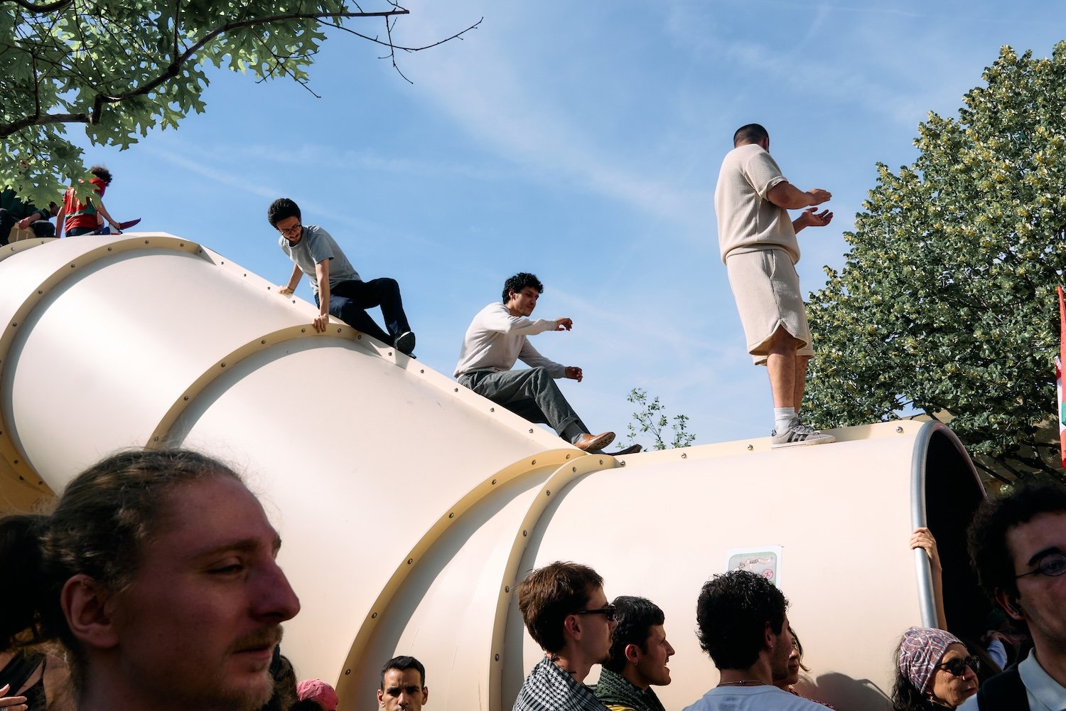 People standing and sitting on a large curved playground slide outdoors, with some climbing and others watching, under a blue sky with trees in the background.