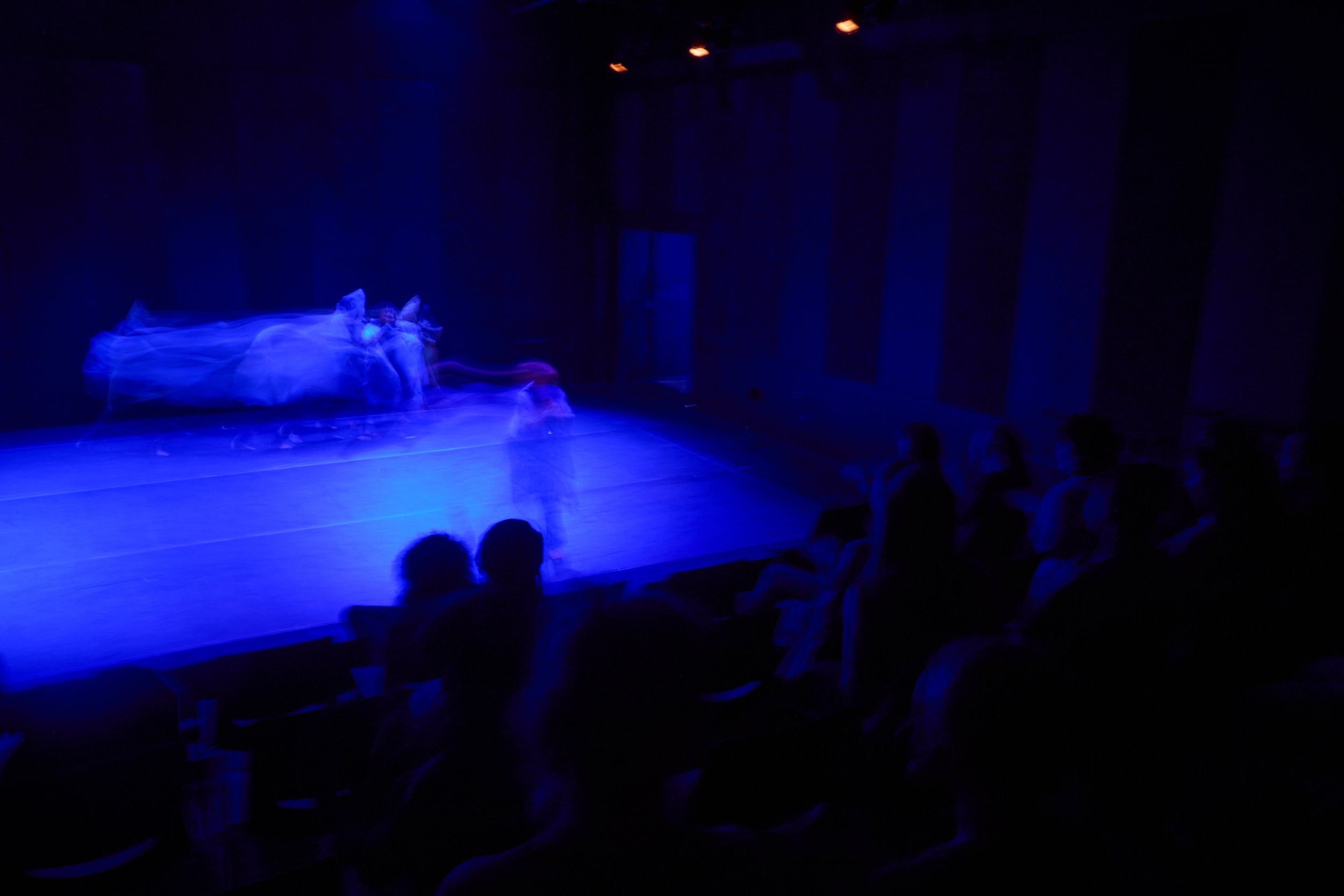 Dark theater with blue stage lighting and performers dressed in white costumes, motion blurred, audience seated in the foreground.