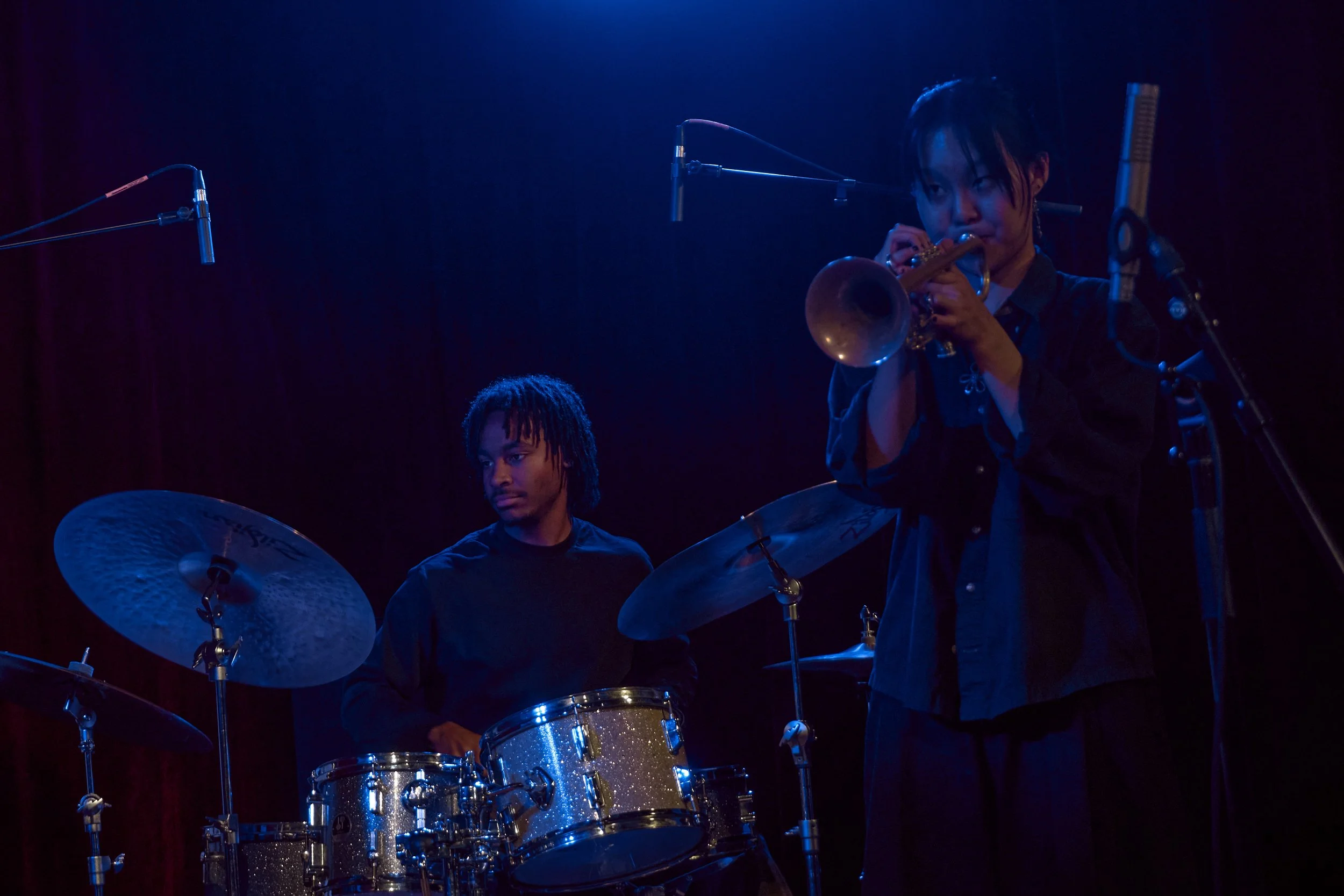 A woman playing the flute and a man playing the drums on stage during a performance, with dark background and stage lighting.